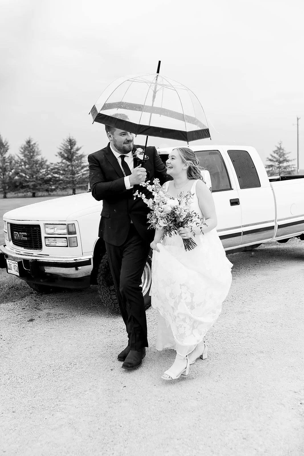 A black and white photo of a newlywed couple walking with an umbrella. The groom is in a suit, holding an umbrella over both, and the bride is in a wedding dress holding a bouquet. They are smiling, and a GMC pickup truck is in the background.