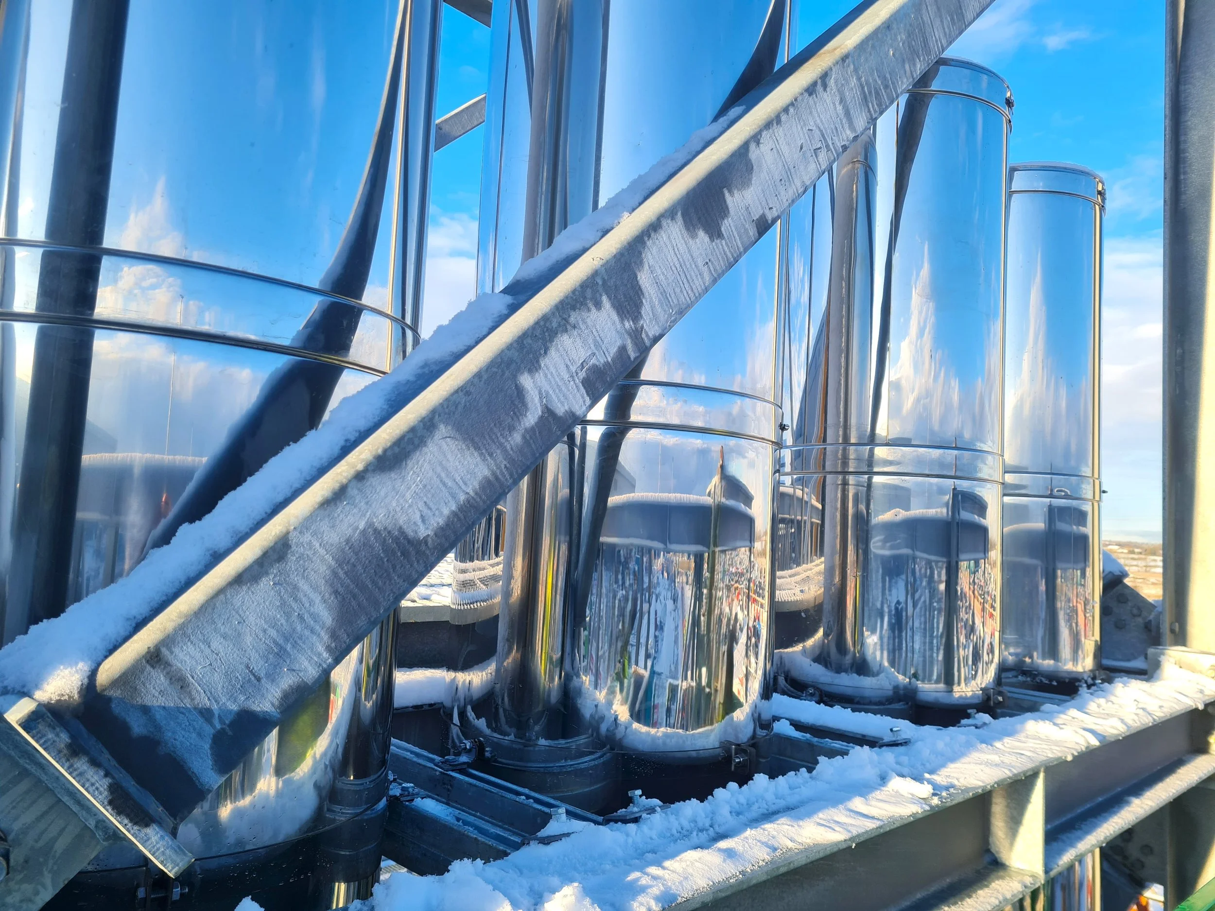 Close‑up of a large twin‑wall stainless steel flue in the snow under a clear blue sky.