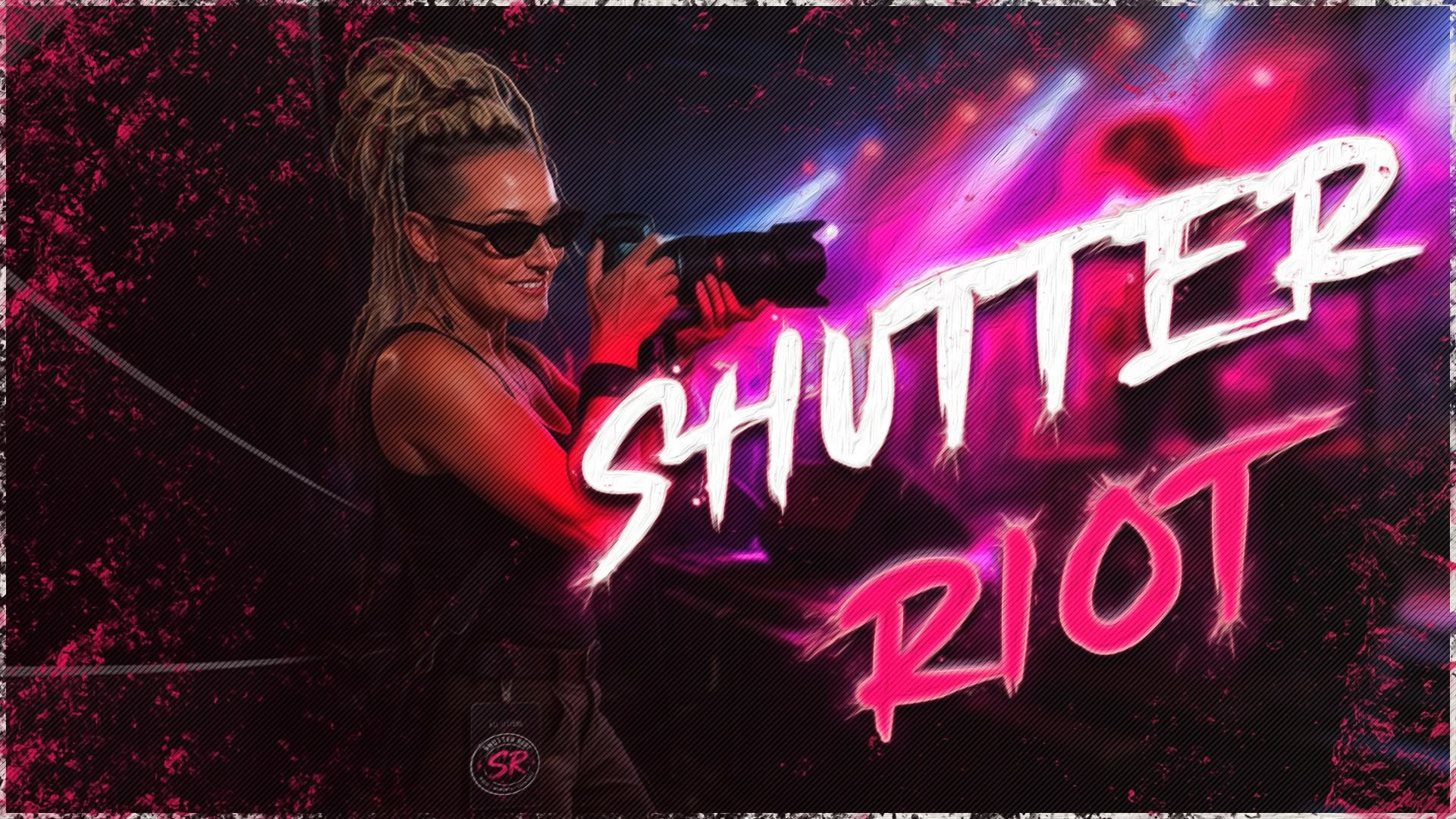 A woman with blonde dreadlocks, wearing sunglasses, holding a camera, standing in front of a neon sign that reads 'SHUT UP 2021' with pink and purple lighting.