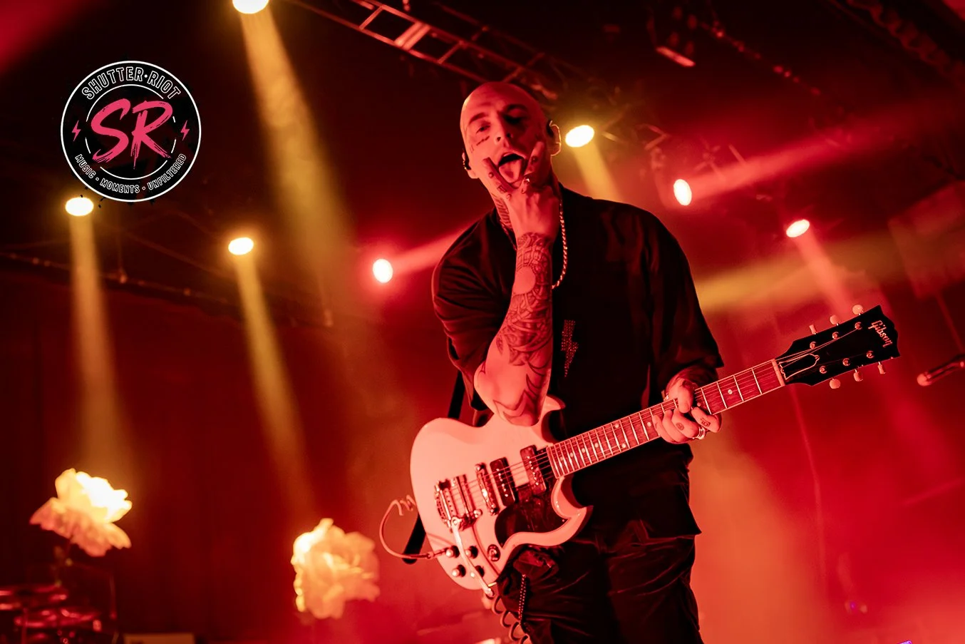A male musician with tattoos on his arm playing a white Gibson electric guitar on stage, illuminated by yellow and red stage lights, with a logo in the top left corner reading 'Shutter Riot SR'.