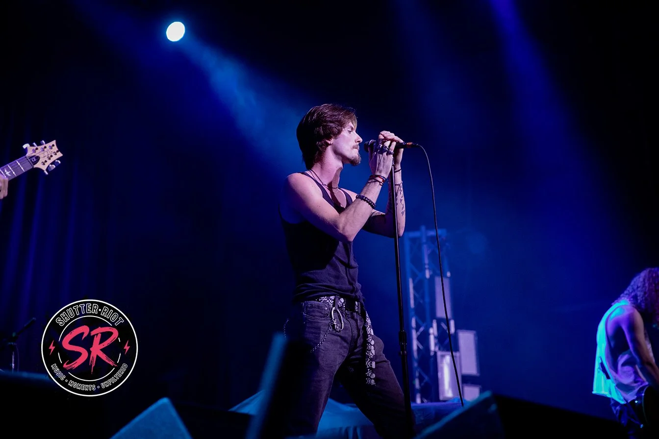 A male singer with dark hair, wearing a sleeveless black shirt and black jeans, performs on stage with a microphone. A blue spotlight shines down from above, illuminating the stage. There is a person to his right and a person to his left, partially visible. The stage background is dark with blue lighting, and a logo with the text "Shutter Riot" is visible in the bottom left corner.
