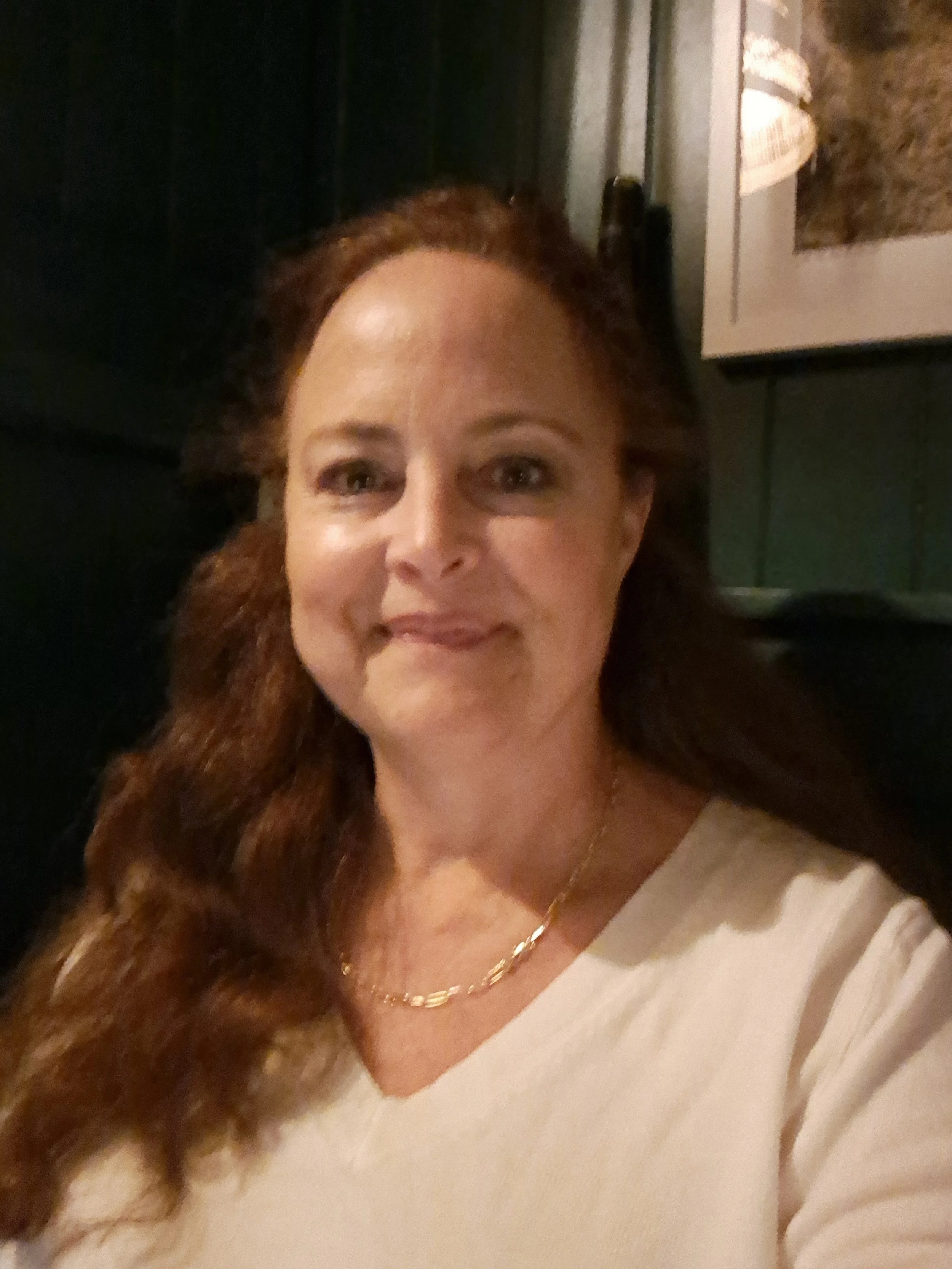 Close-up of a woman with ginger hair, wearing a white top and a gold necklace, smiling in a dimly lit room.