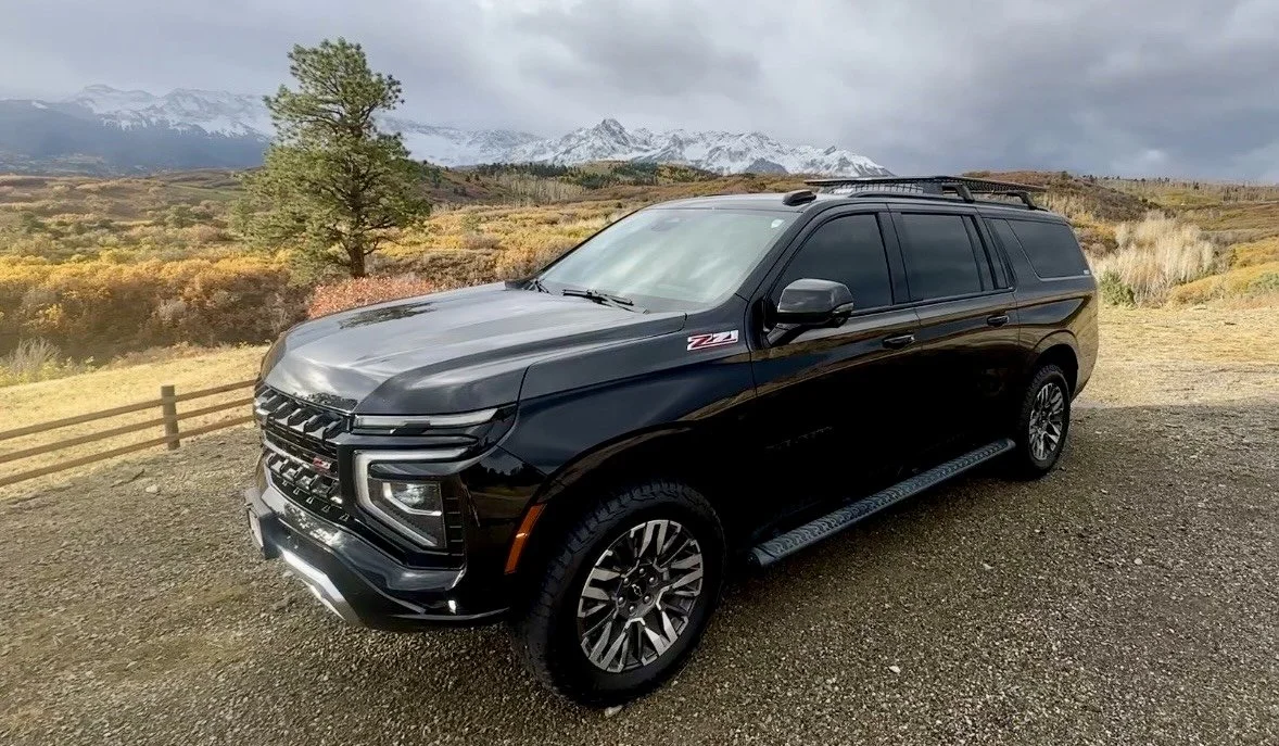 A new black Chevy Suburban Z71 parked at the summit of a mountain pass near Telluride, Colorado, surrounded by vibrant golden autumn foliage and backed by towering snow-capped peaks.