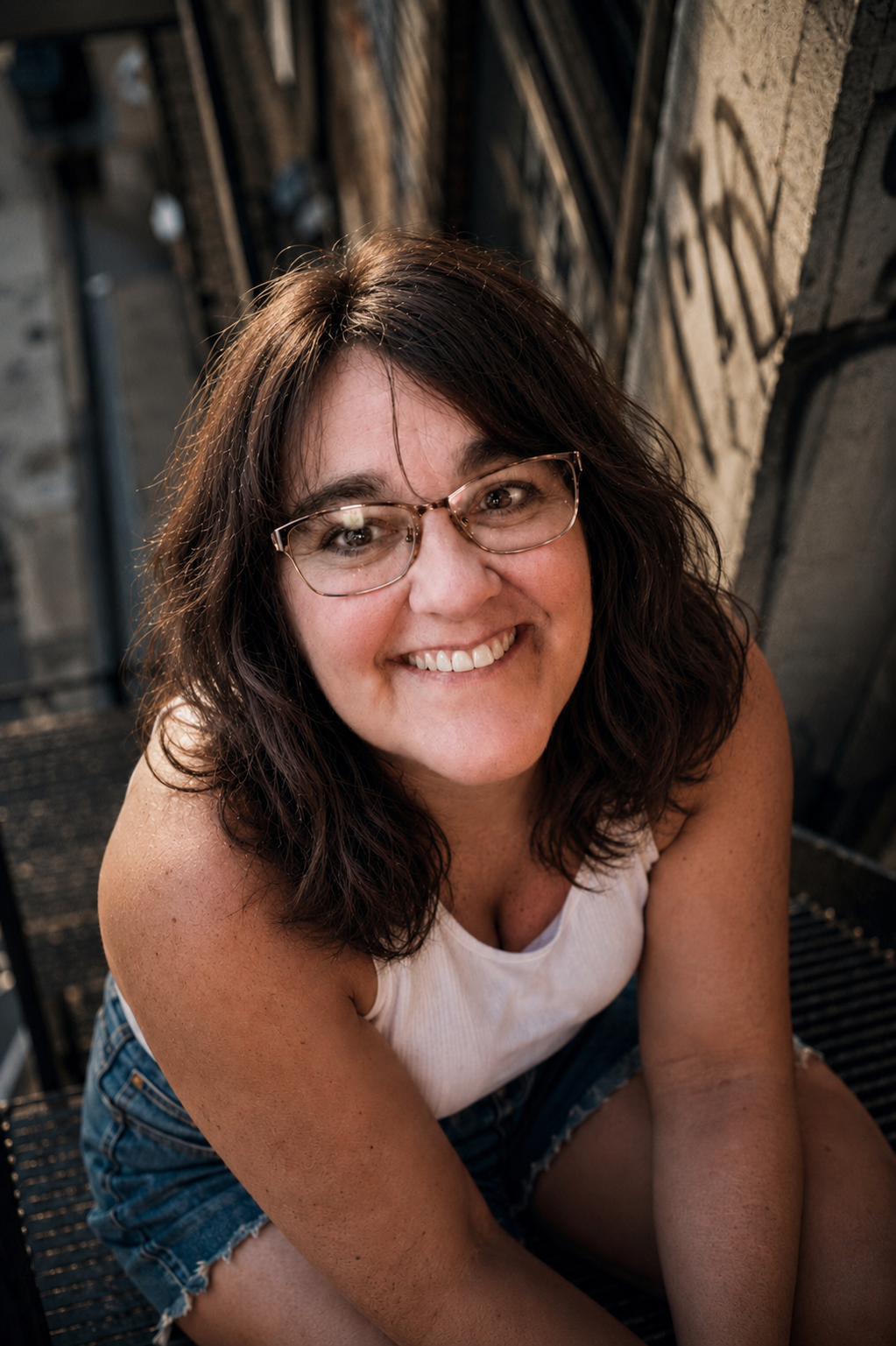 A woman with shoulder-length dark hair, glasses, and a white tank top smiling while sitting outdoors on a metal staircase, with brick and metal background.