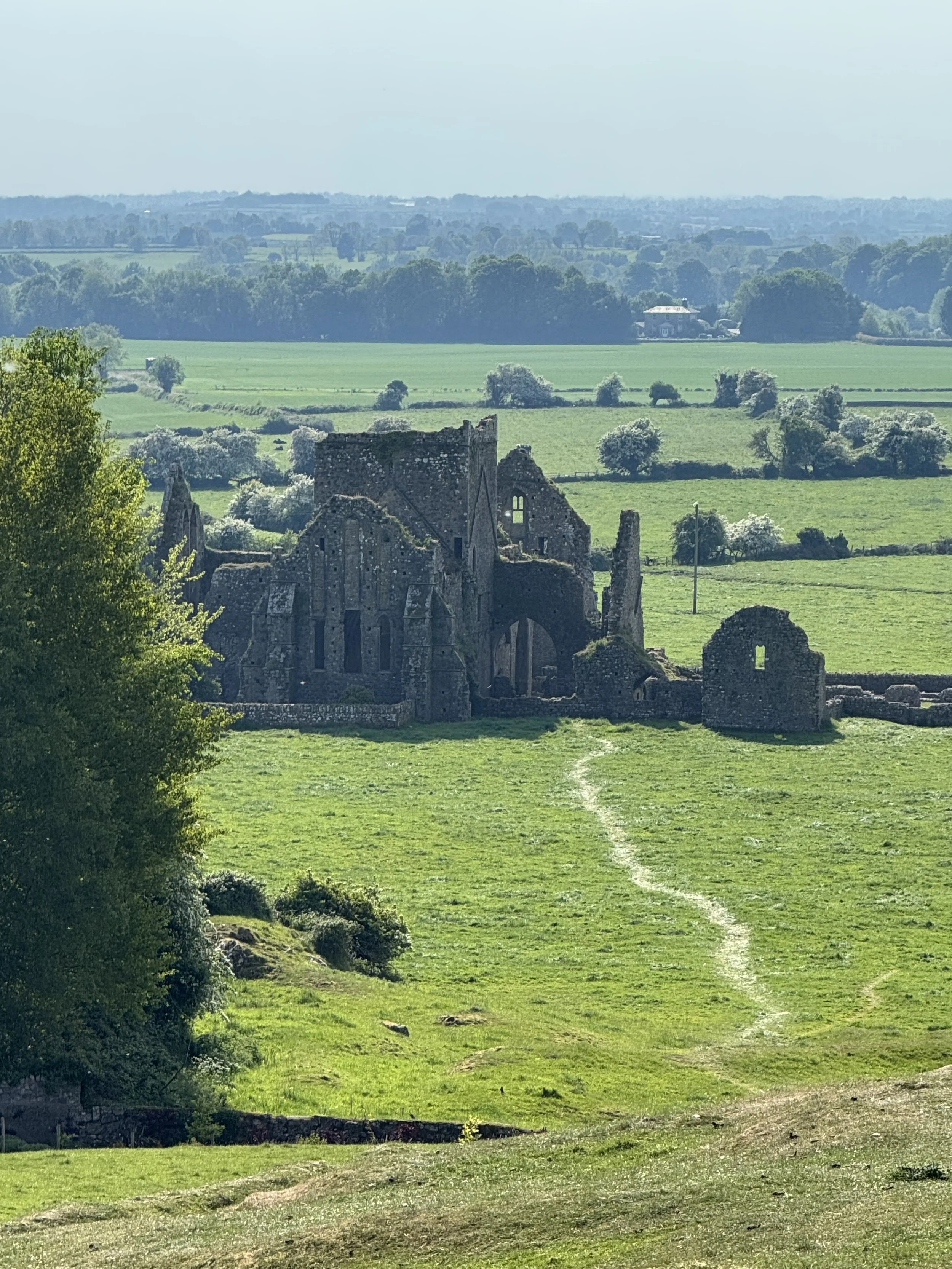 Ruins of an old stone church in a lush green landscape with trees and fields, under a hazy sky.