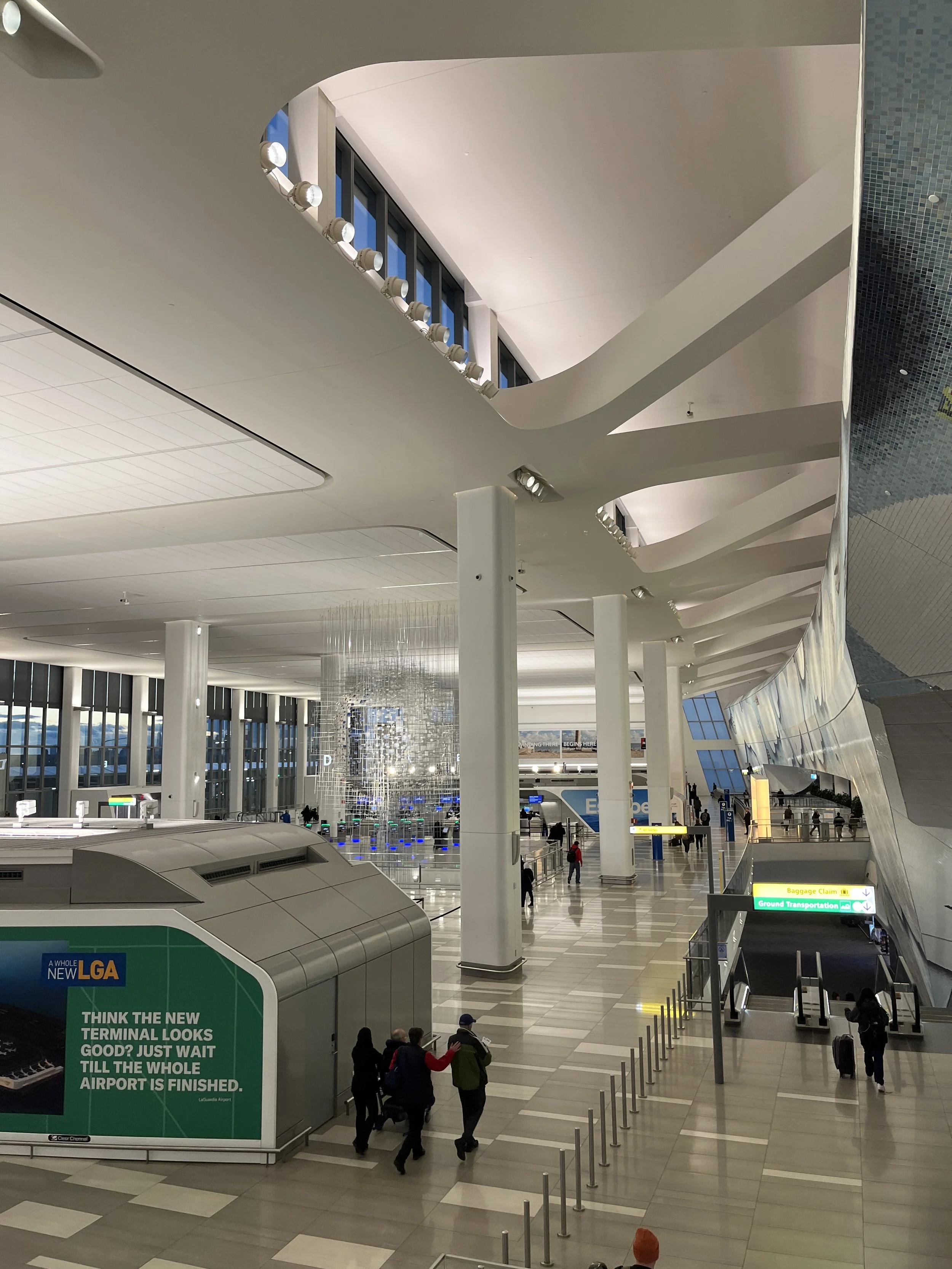 Interior of an airport terminal with people walking through luggage claim area, escalators, and signage for baggage claim and ground transportation.
