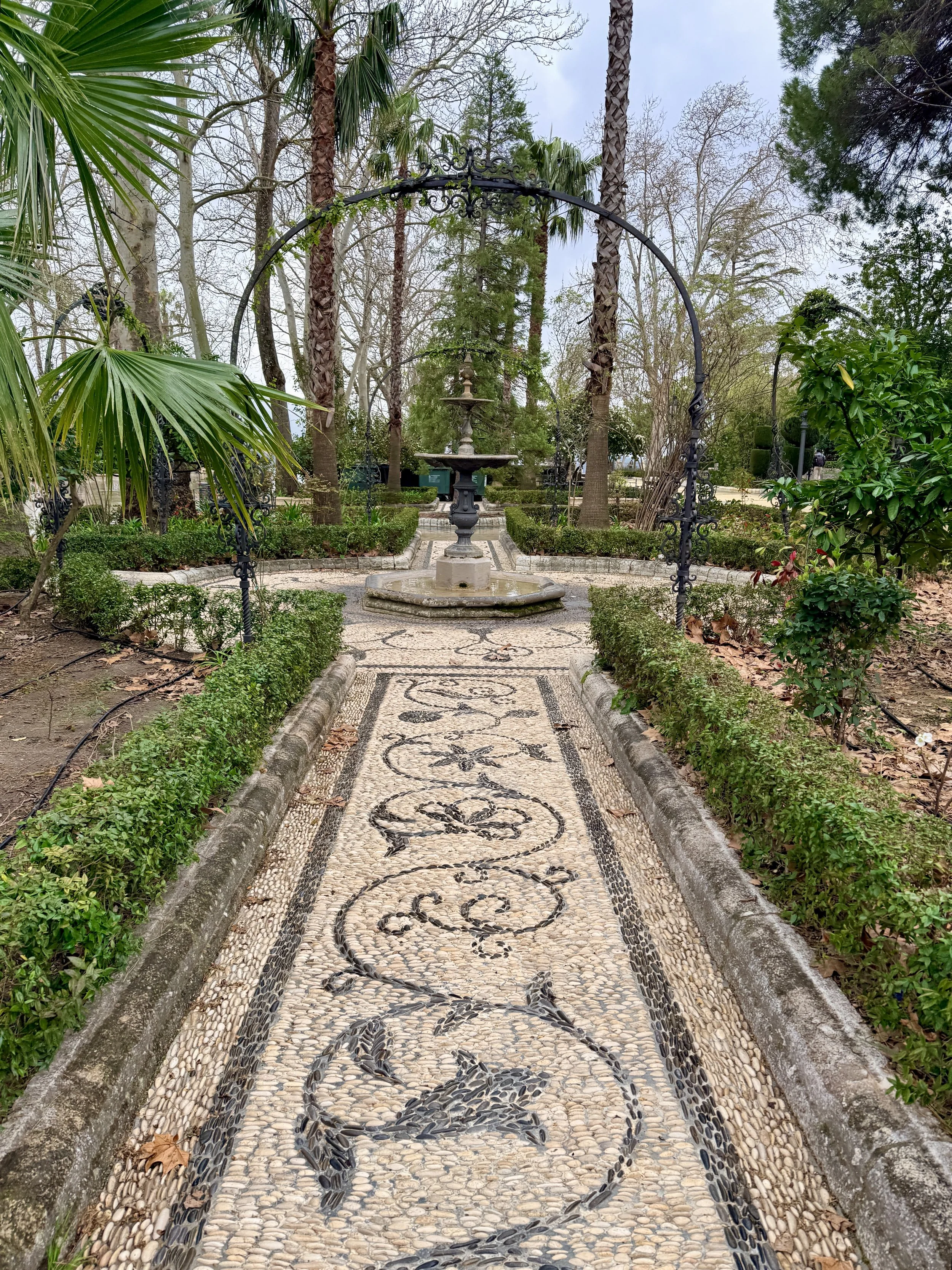 A stone pathway with intricate black pebbled designs of swirling patterns, bordered by low hedges, leading to a fountain with a multi-tiered structure in a lush garden surrounded by tall trees.