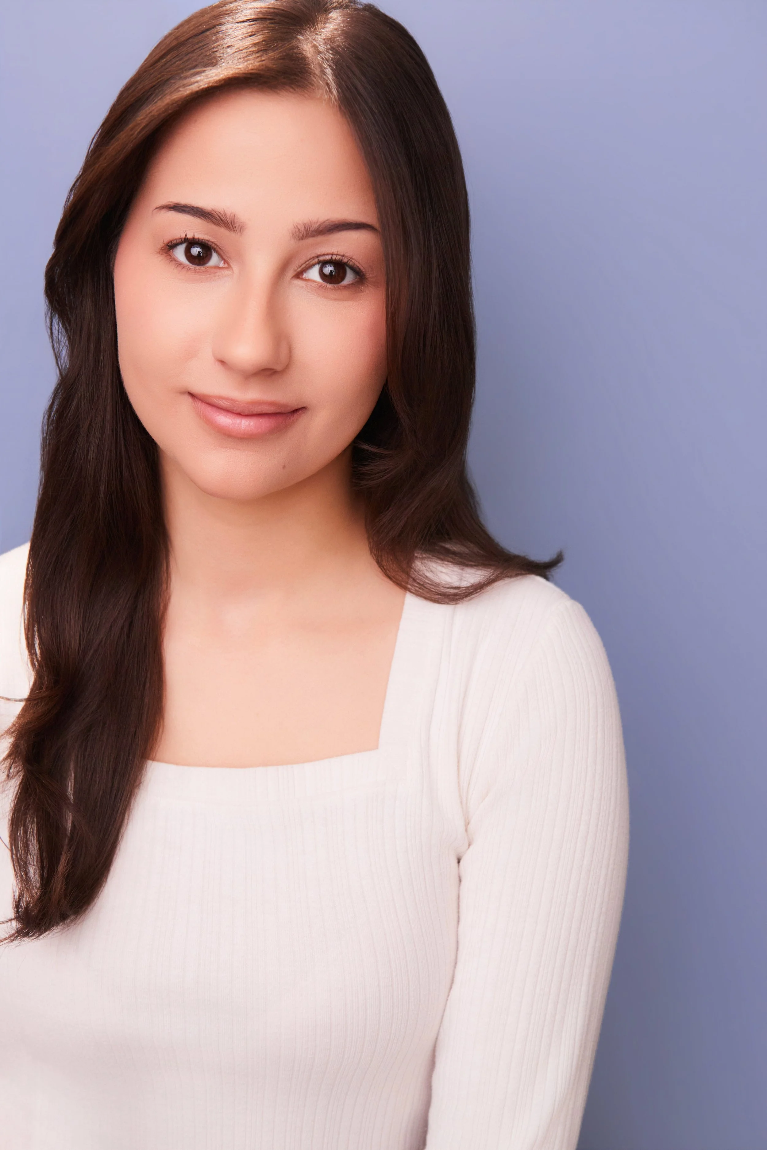 Close-up portrait of a smiling woman with long brown hair wearing a white top against a lavender background.