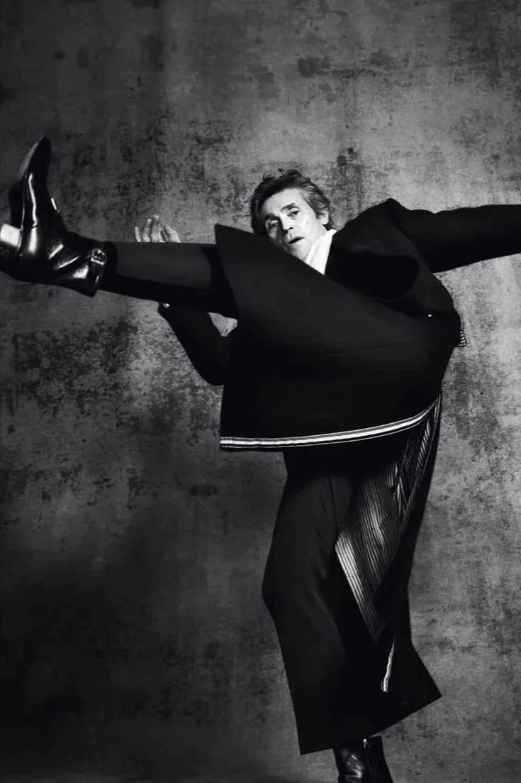 Black-and-white photo of a man in a suit performing a high kick in front of a textured backdrop.
