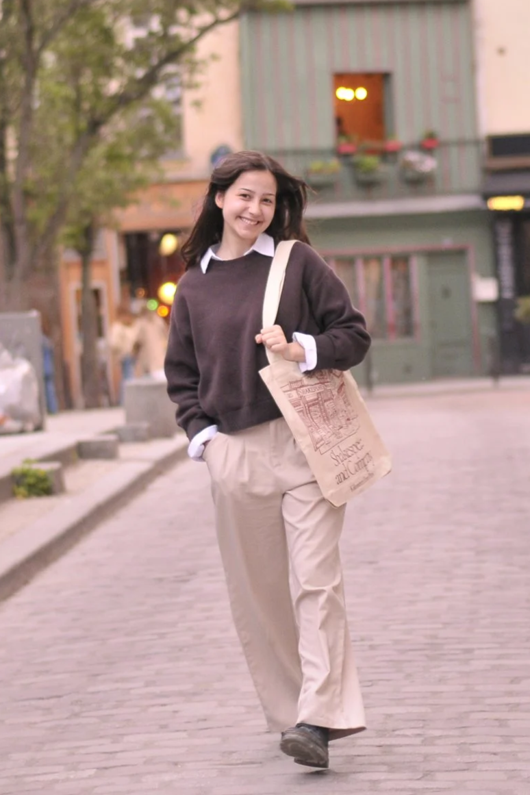 A young woman smiling and walking on a city street at sunset, holding a beige "Shakespeare & Co." tote bag.