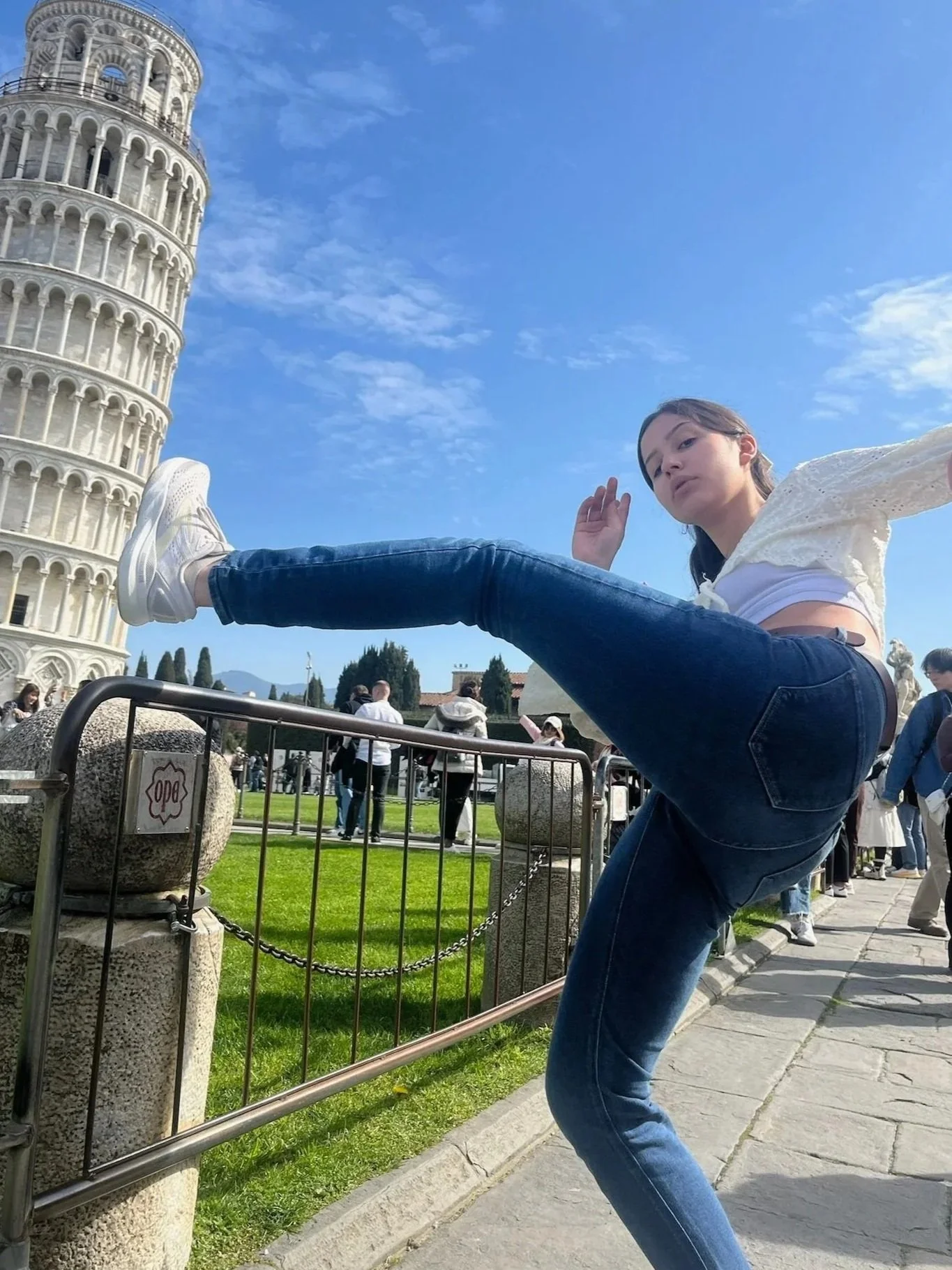 A woman posing with one leg raised on a railing near the Leaning Tower of Pisa. She is wearing a white top, blue jeans, and white sneakers, with a clear sky and other tourists in the background.