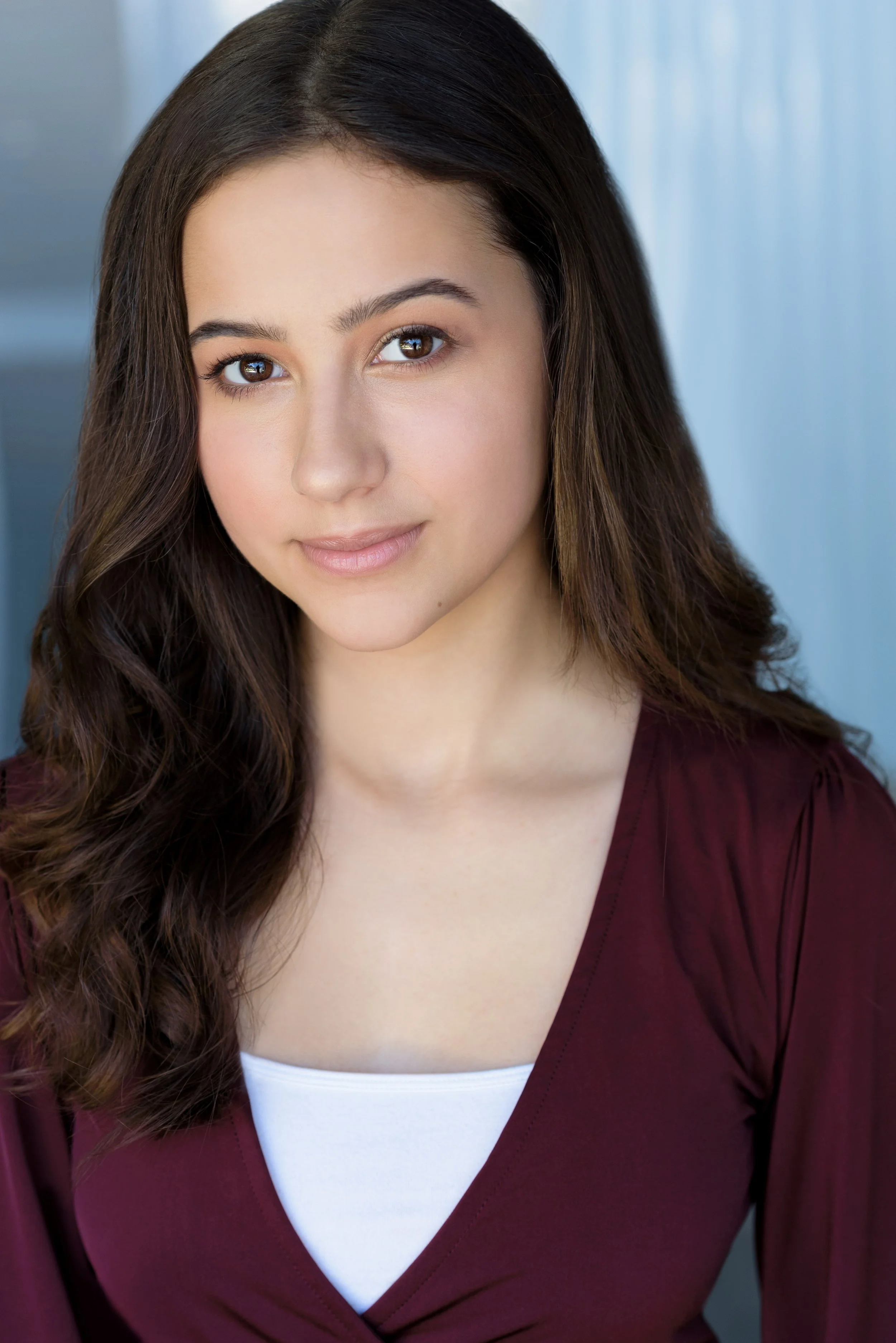 Close-up of a young woman with long, dark wavy hair, brown eyes, and a light complexion, wearing a maroon top and white undershirt, standing against a blue-toned background.