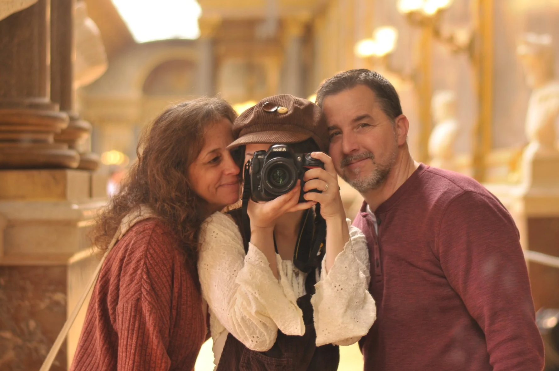 Three people taking a selfie together inside a grand, ornately decorated building. The person in the middle is holding a camera, and all three are close together, smiling, with the couple on either side leaning in towards the person with the camera.