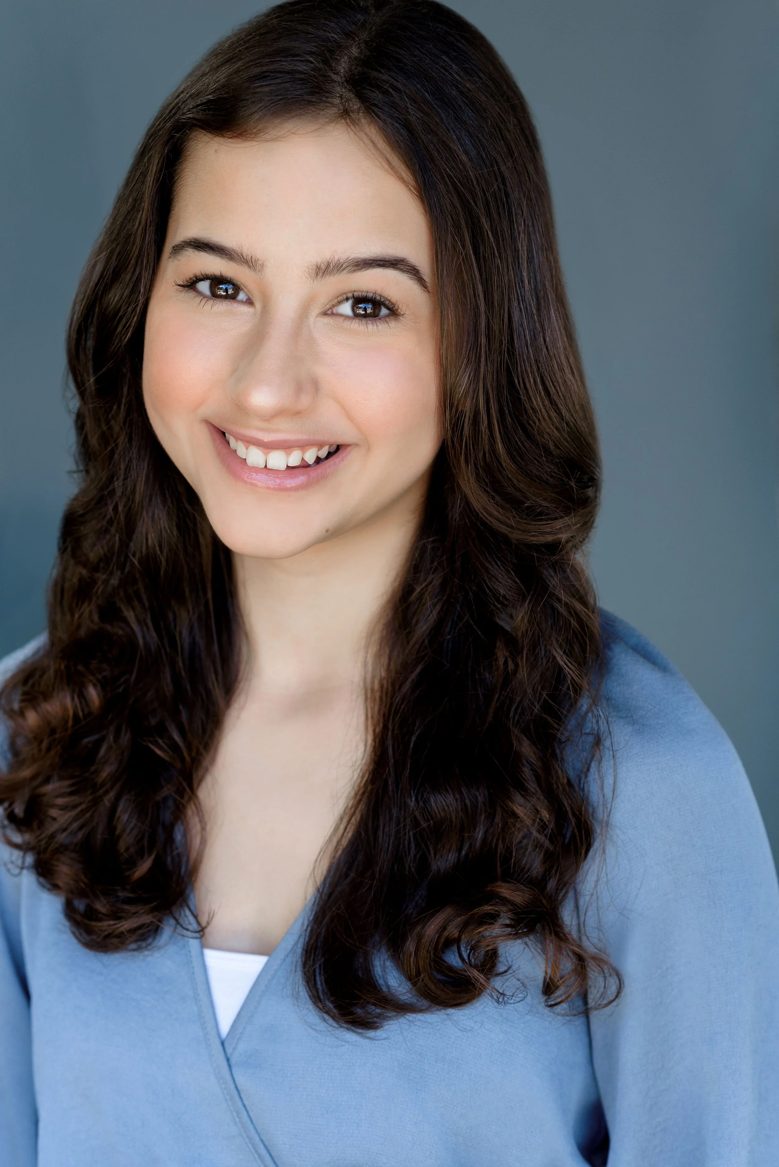 A young woman with long, wavy brown hair smiling, wearing a light blue top, and standing against a solid blue-gray background.