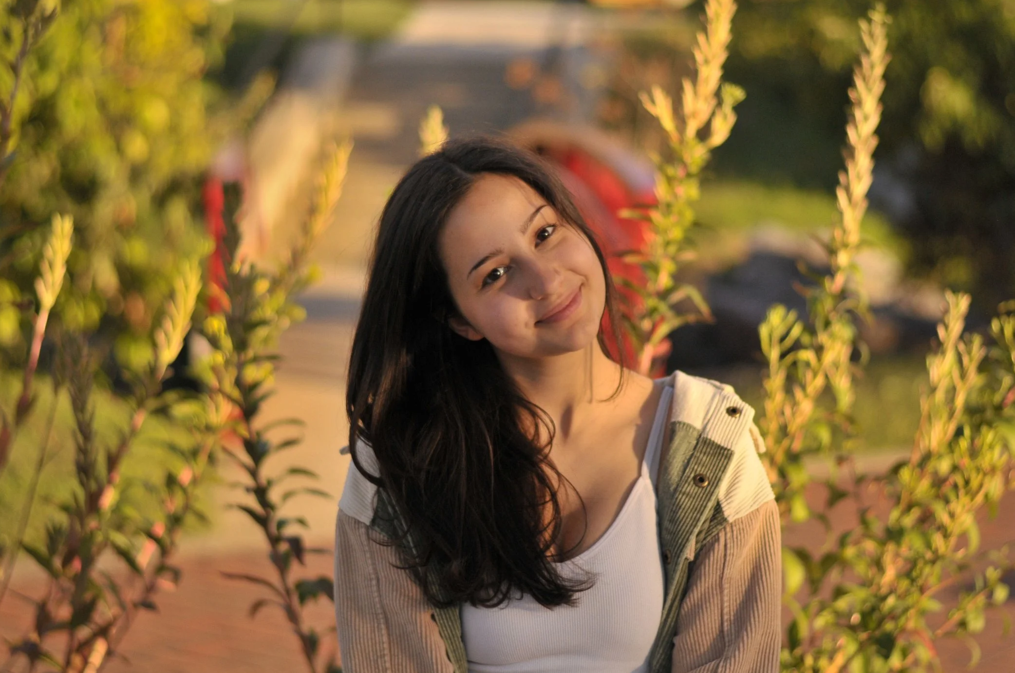 A young woman with long dark hair smiling, standing among green plants outdoors during sunset.