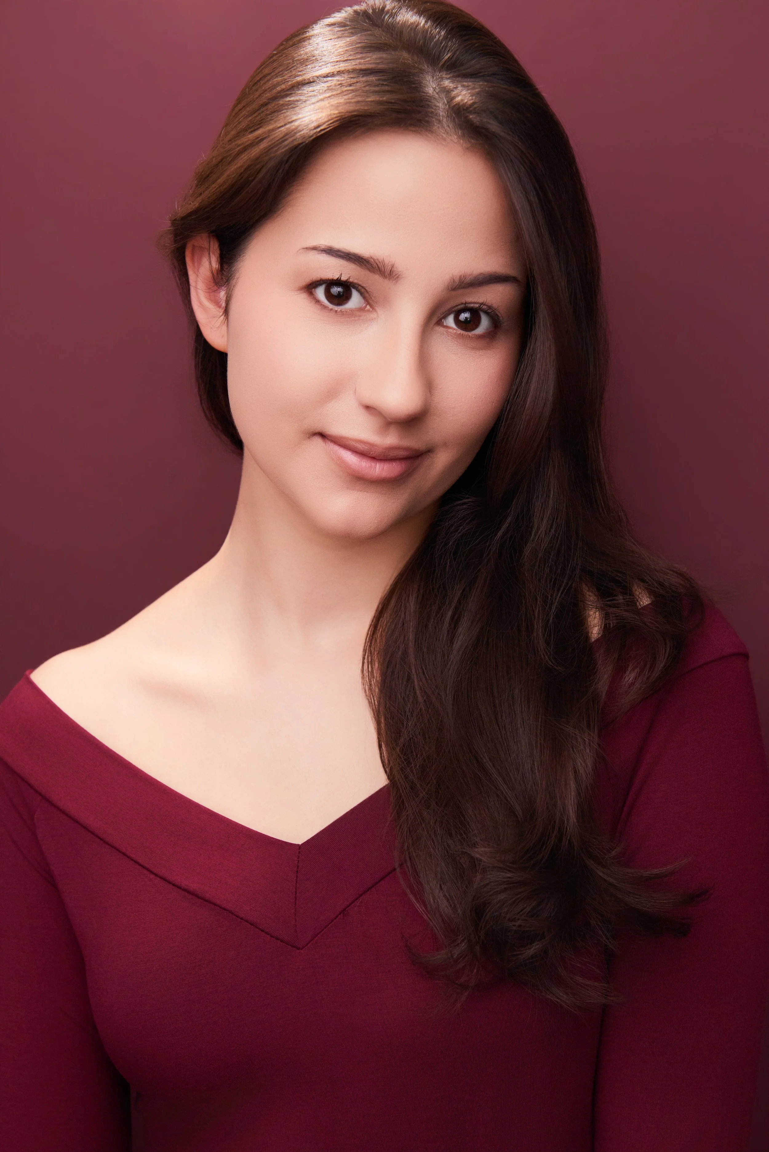 A woman with long, dark, wavy hair styled over her right shoulder, wearing a maroon, off-the-shoulder top, smiling subtly against a solid mauve background.