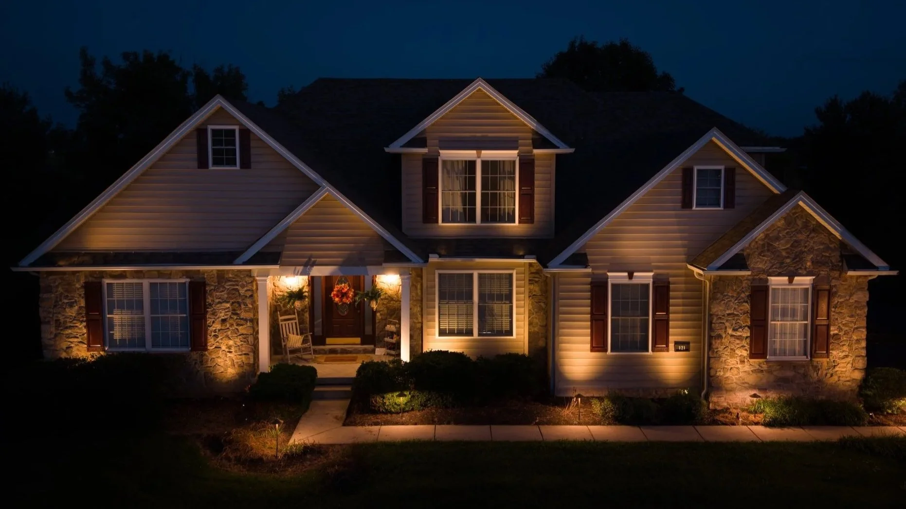 Large suburban house with stone accents, warm exterior lighting, and landscaped front yard at night