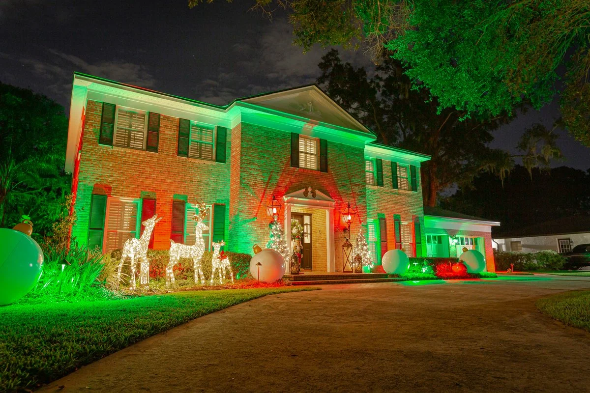 Brick two-story home decorated with green and red Christmas lights, illuminated reindeer, ornaments, and wreaths at night