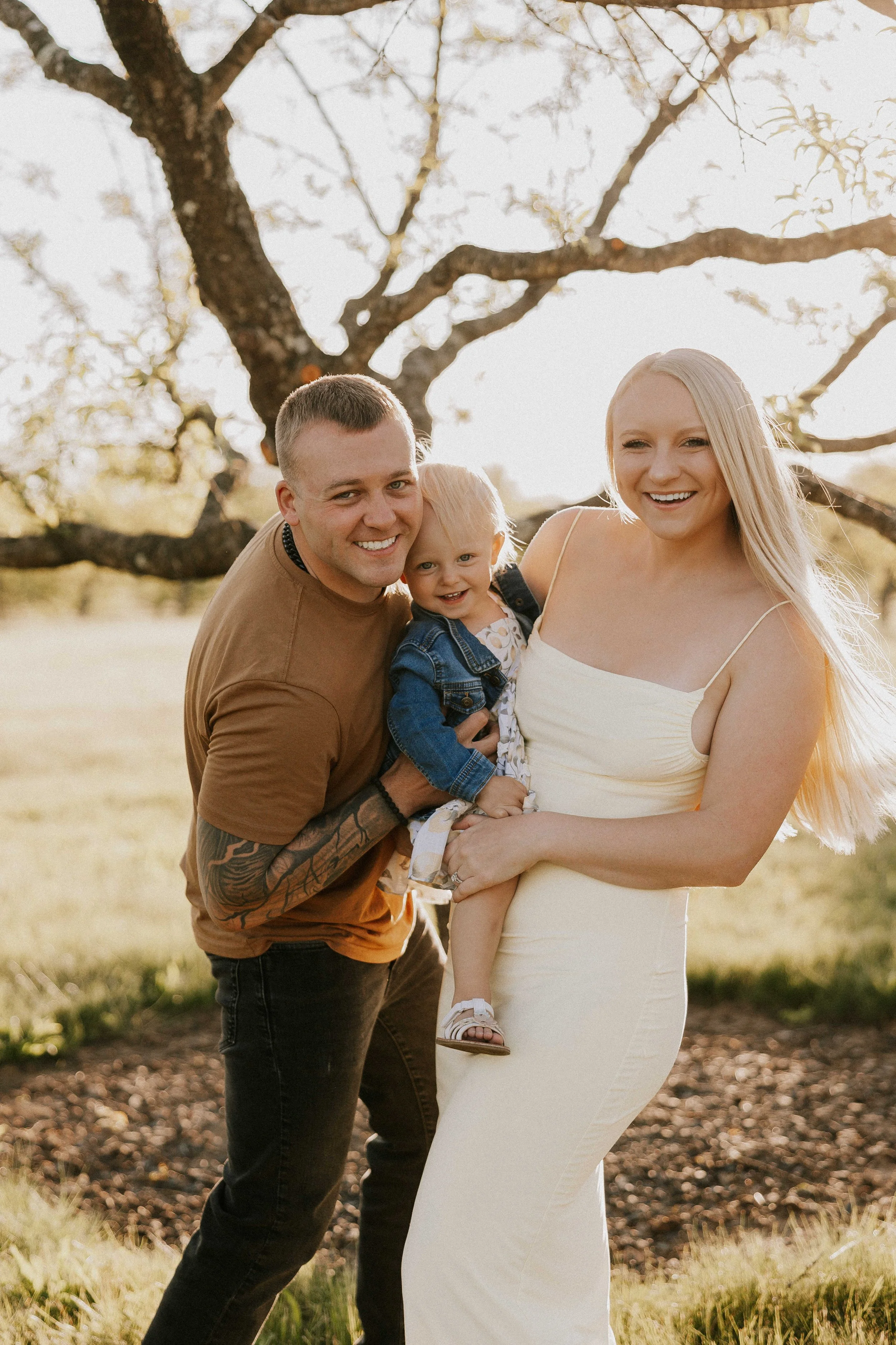 Smiling family of four posing outdoors beneath a flowering tree, with parents holding their young child.