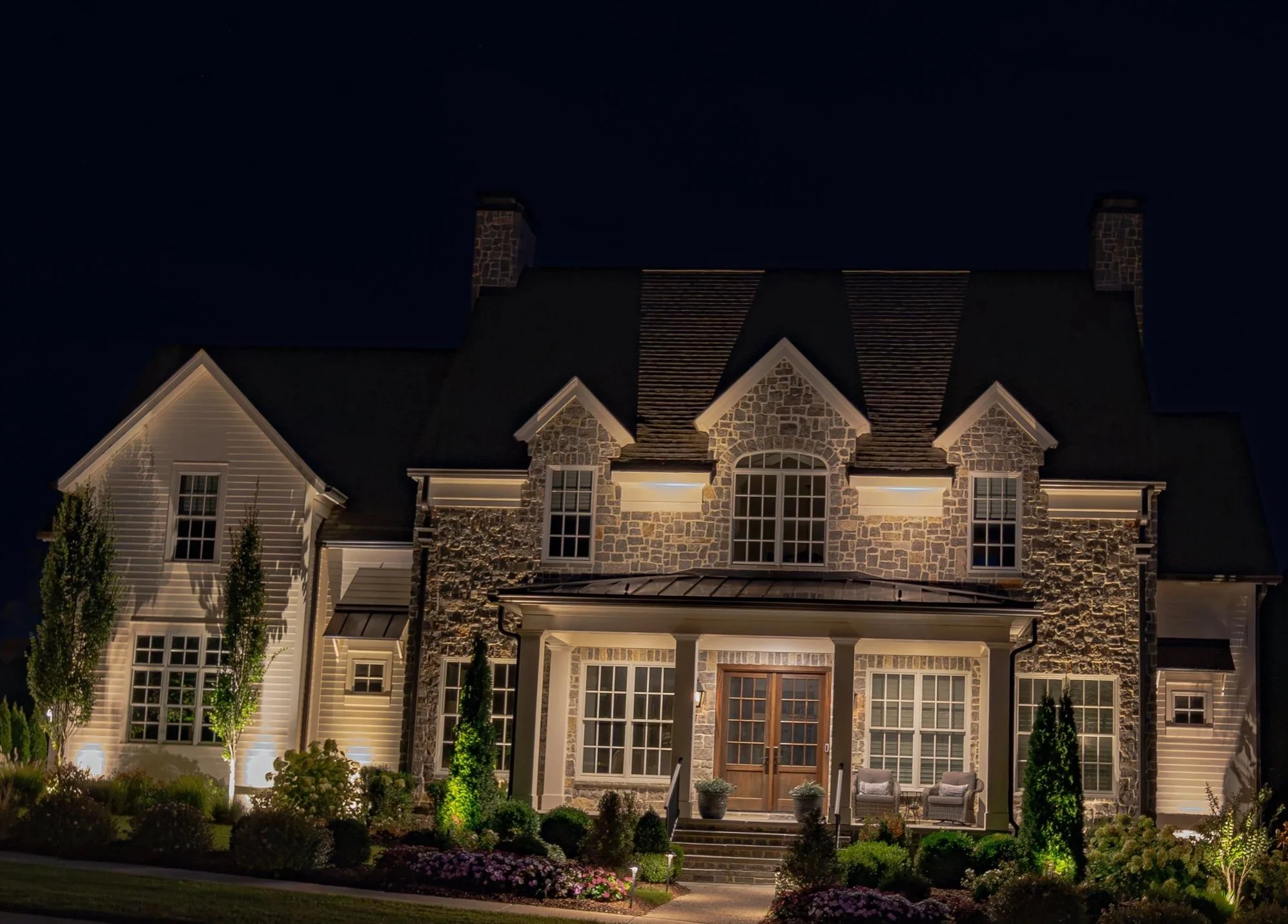 Large illuminated stone-and-siding luxury home with landscaped front yard at night