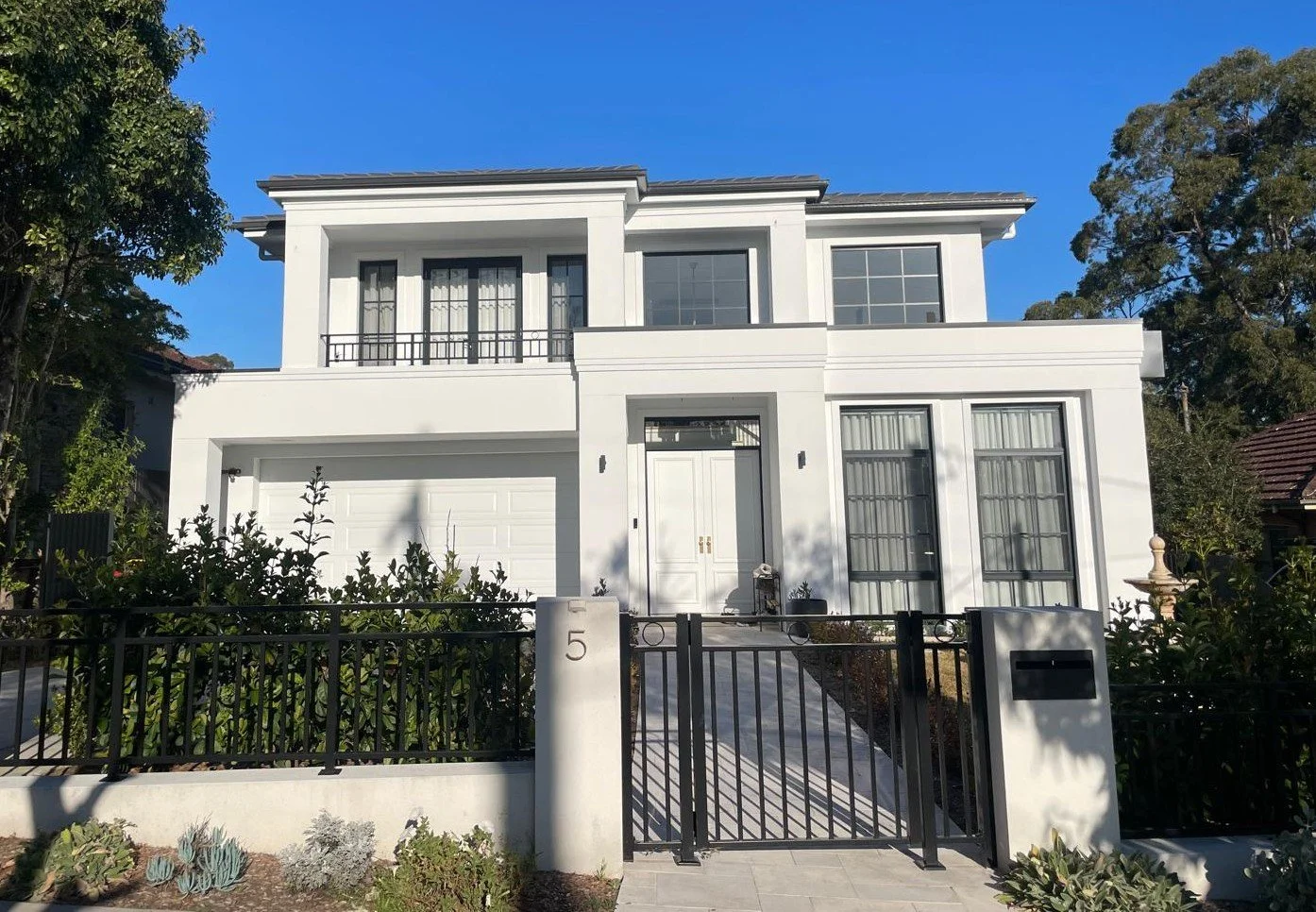 Modern white two-story house with a black gate and a small front garden, blue sky, and some trees in the background.
