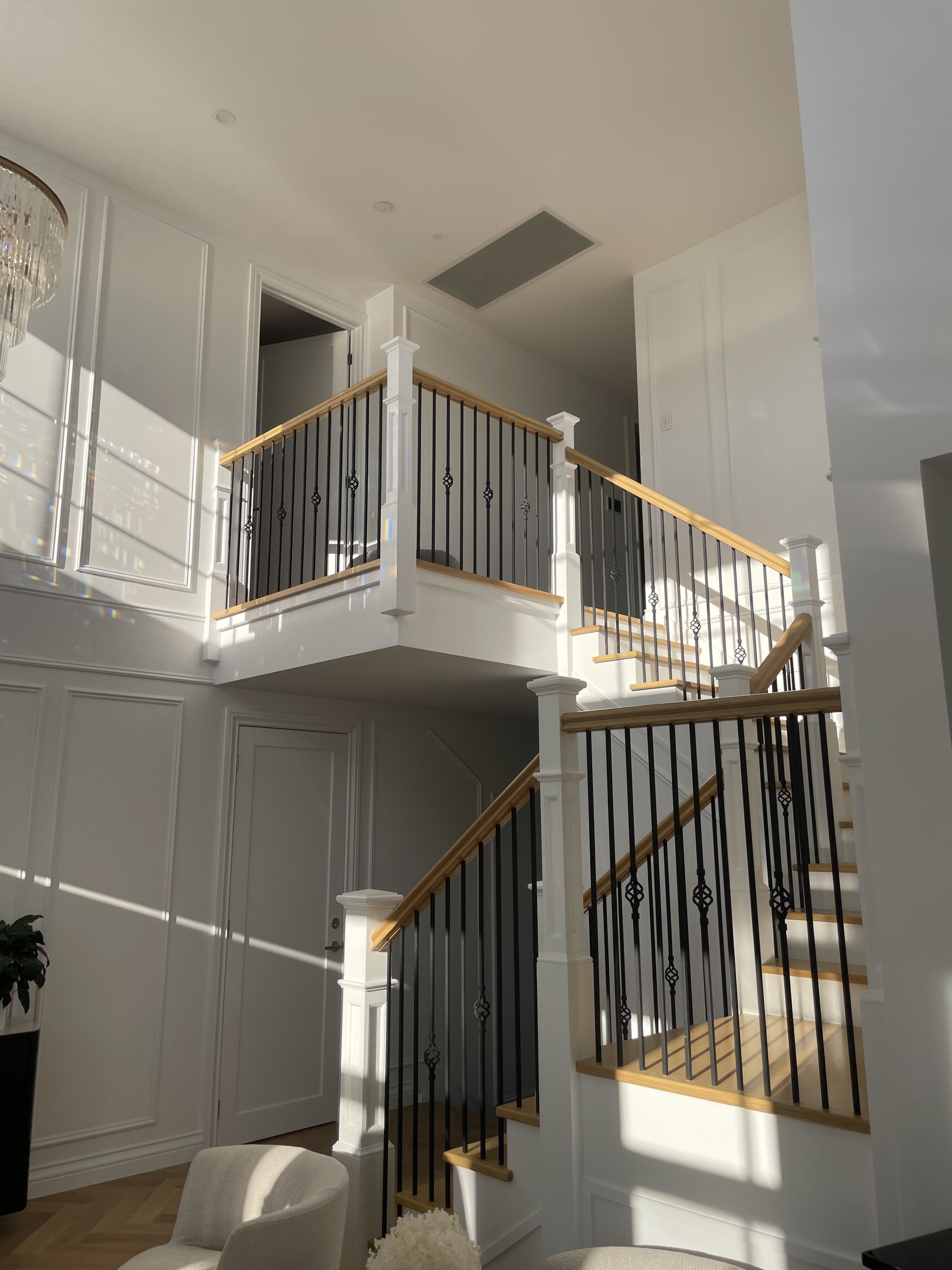 Interior view of a modern staircase with wooden steps and black metal railing, white walls with decorative paneling, and sunlight casting shadows on the wall.