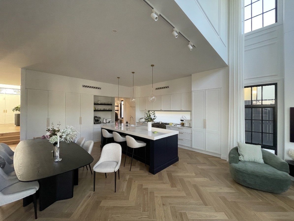 Bright, modern kitchen and dining area with white walls and cabinets, black island, and natural wood herringbone flooring. Features pendant lights, large windows, and a green lounge chair.