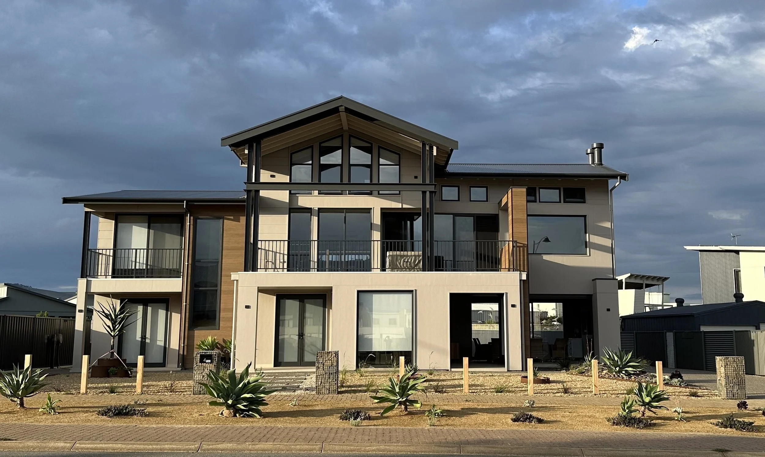 A modern multi-story house with large windows, wooden accents, and a balcony, with a landscaped front yard and cloudy sky in the background.