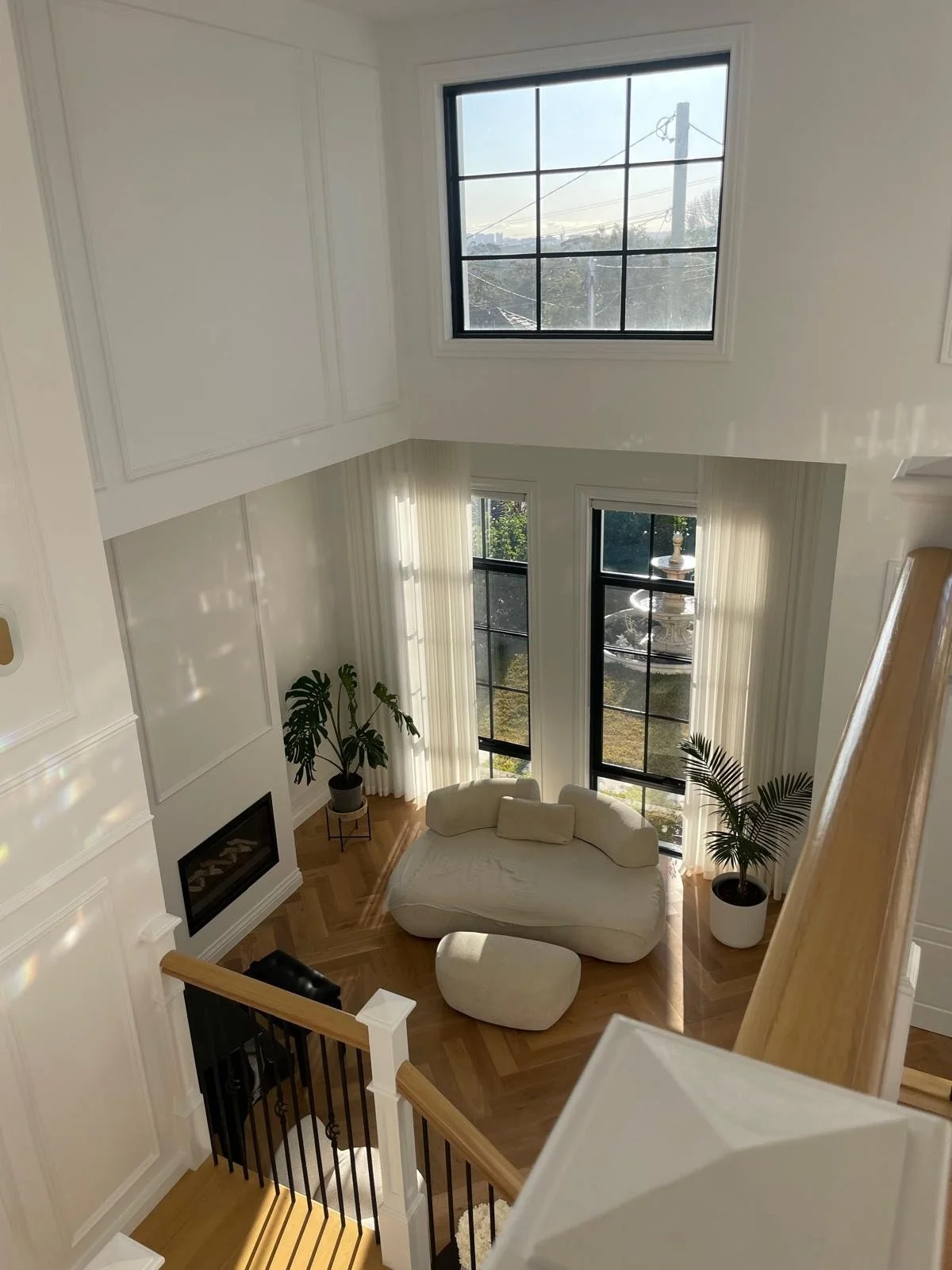 The image shows a living room with high ceilings, large windows, a white modern sofa, two plants in white pots, a white curtain, and a fireplace with a wooden floor.