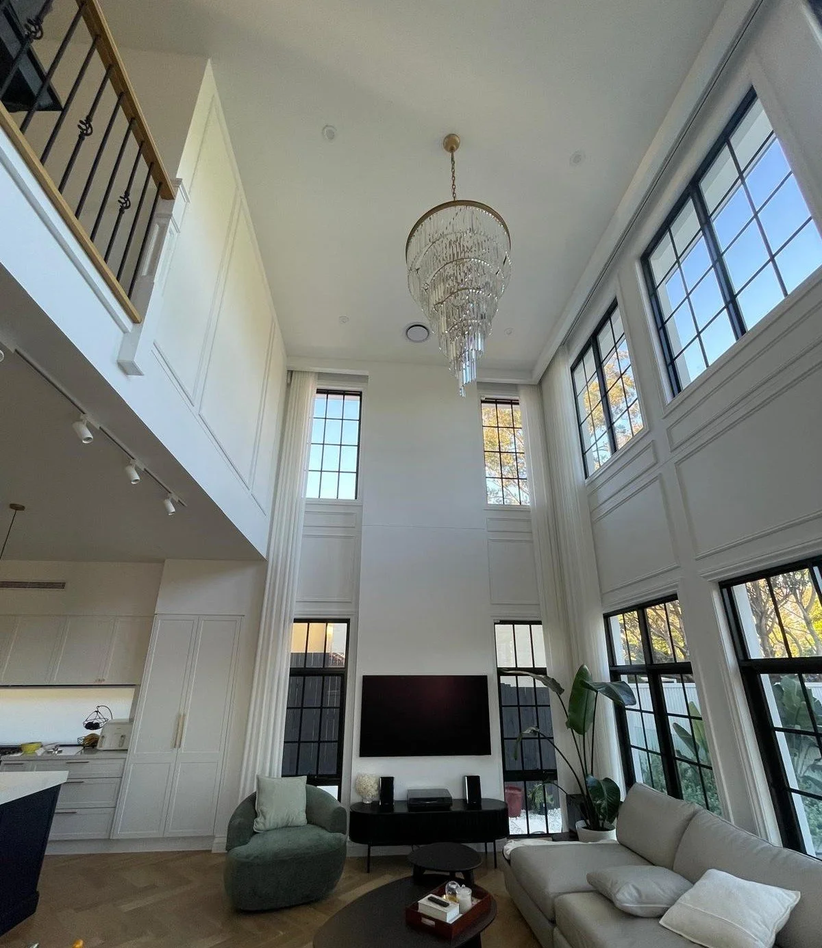 Living room with high ceiling, large windows, a chandelier, TV, modern furniture, and a kitchen partially visible.
