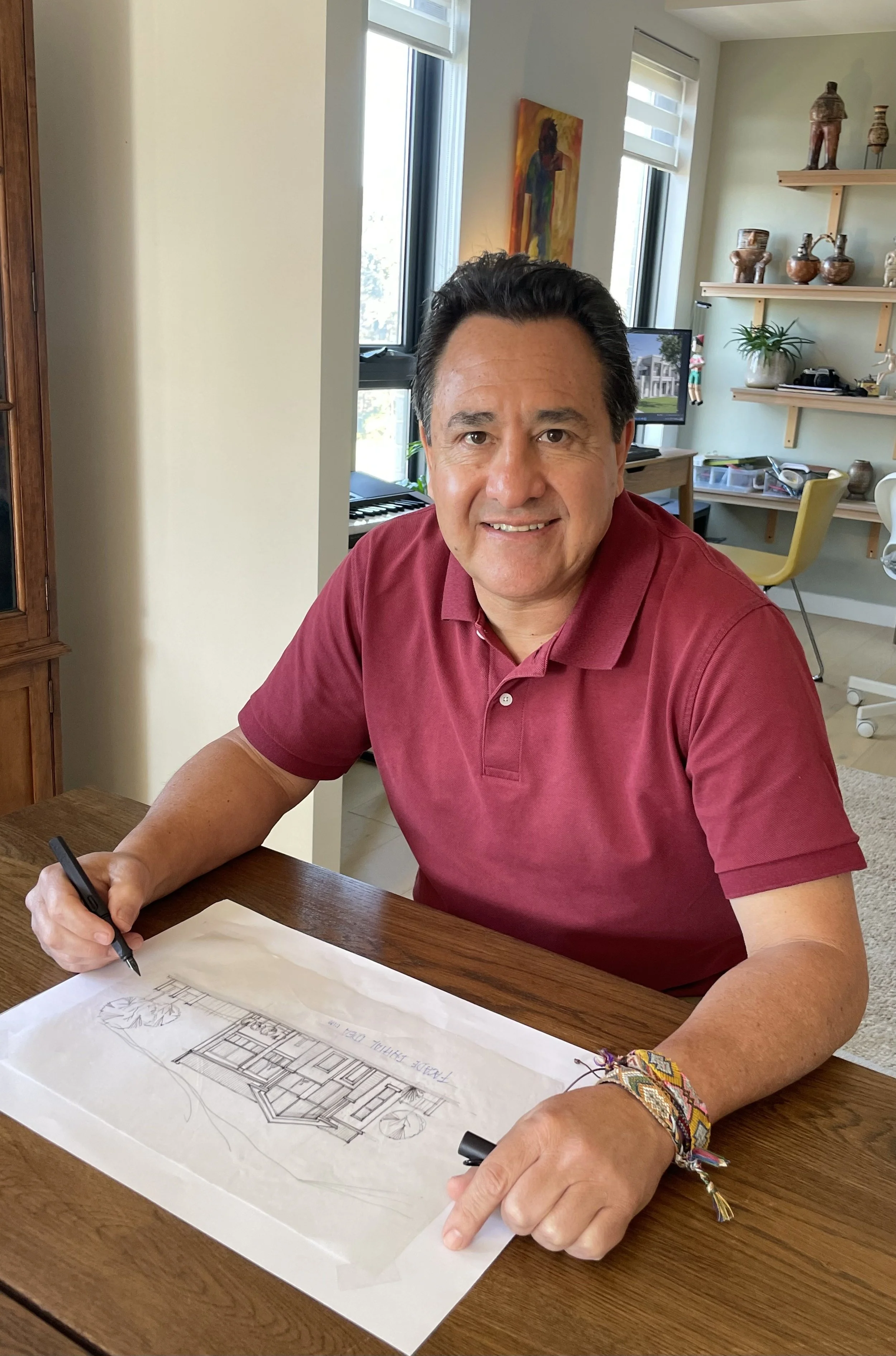 A man in a red polo shirt sitting at a wooden table, drawing architectural plans on paper with a black pen. He has dark hair and is wearing colorful bracelets.