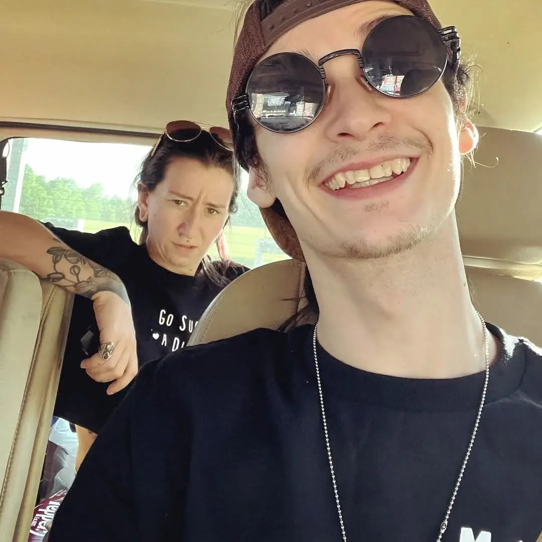 Two young adults riding in a vehicle, taking a selfie. The man in the foreground is smiling, wearing dark sunglasses and a brown hat. The woman in the background has a serious expression, sunglasses on her head, and tattoos on her arm.