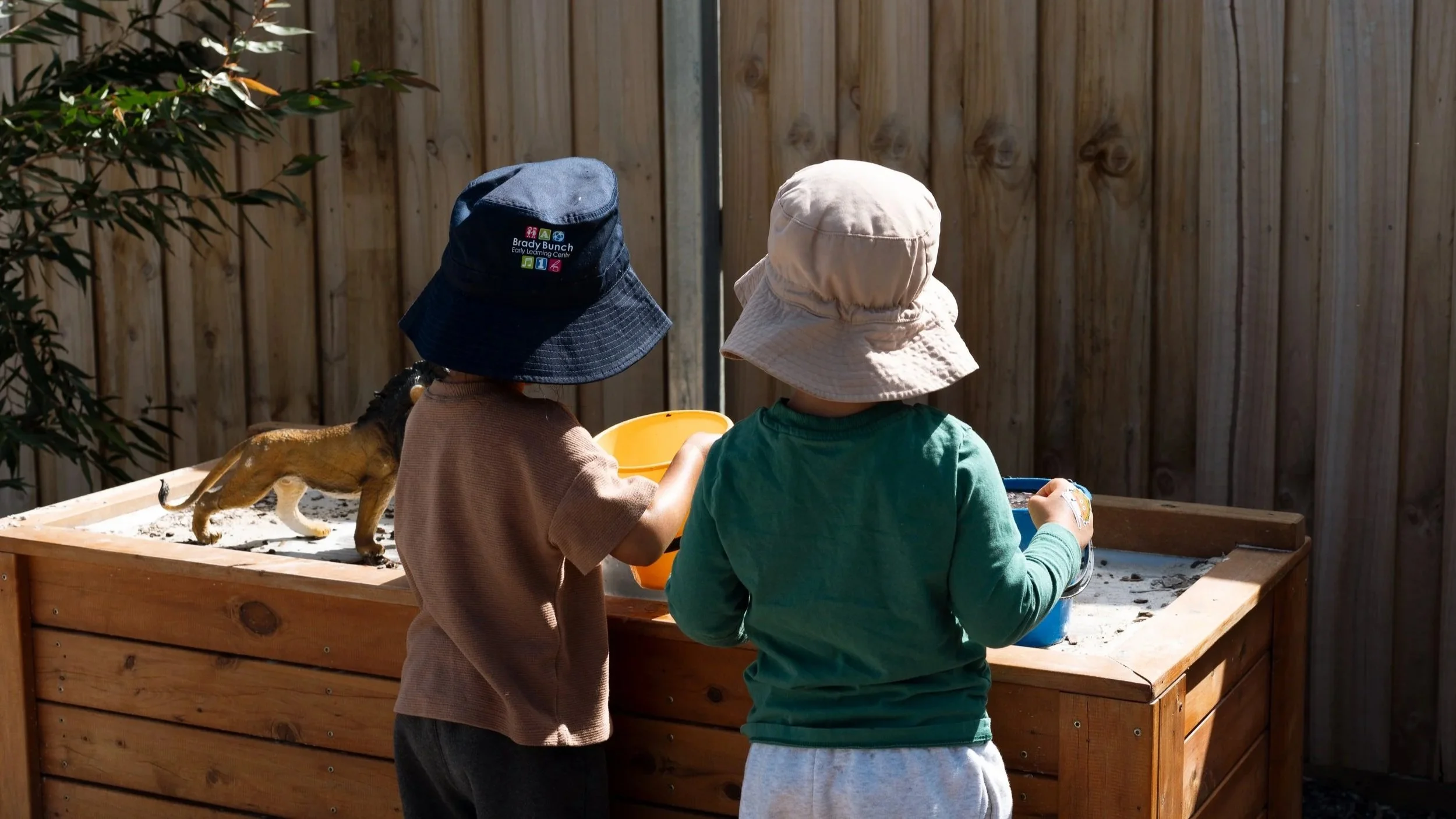 Two children wearing hats and long-sleeved shirts feeding a lion cub in a wooden enclosure, with a wooden fence in the background.