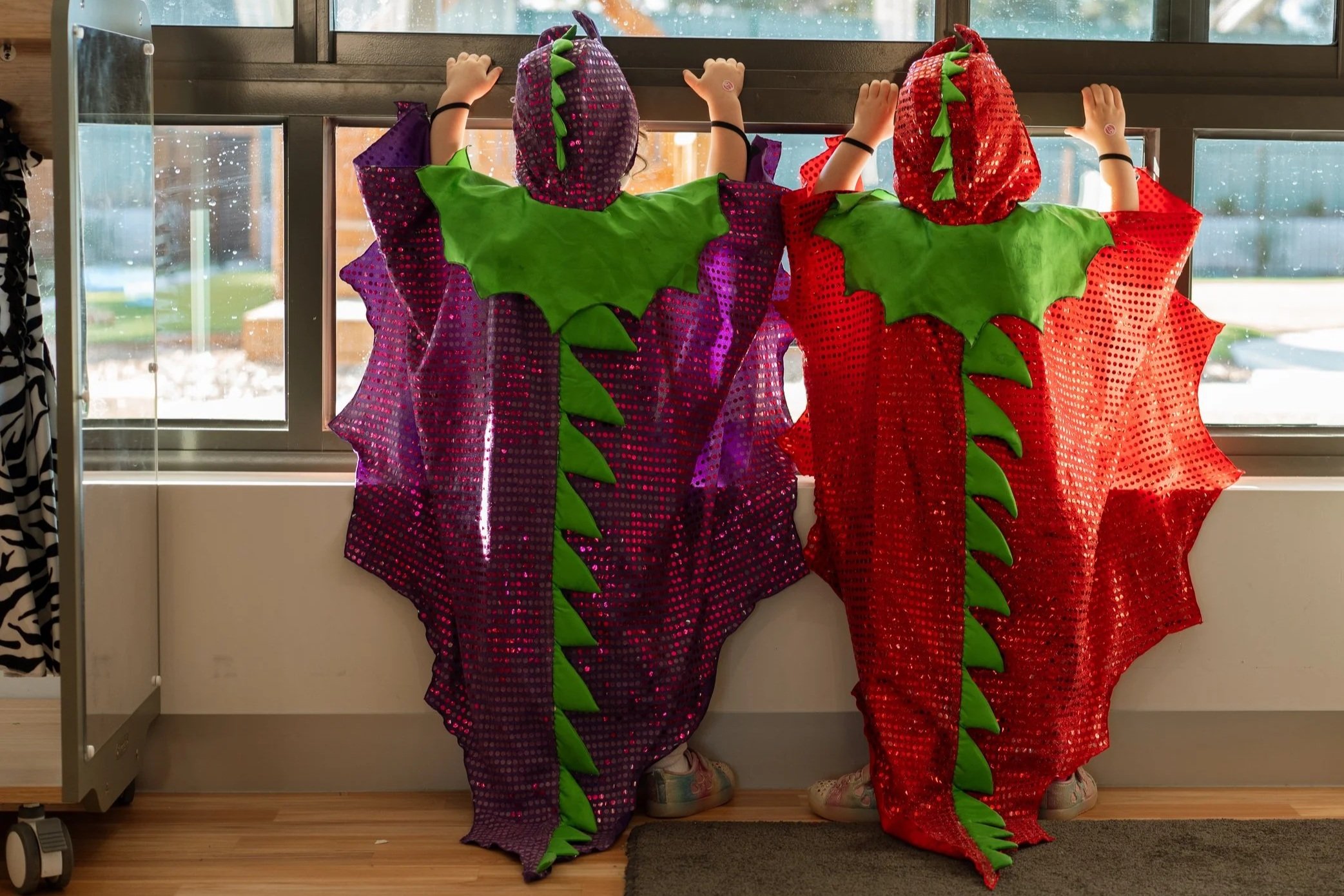 Two children dressed in colorful dragon costumes made of shiny fabric with green scales, standing by a window with rain streaks visible, holding onto the window frame.