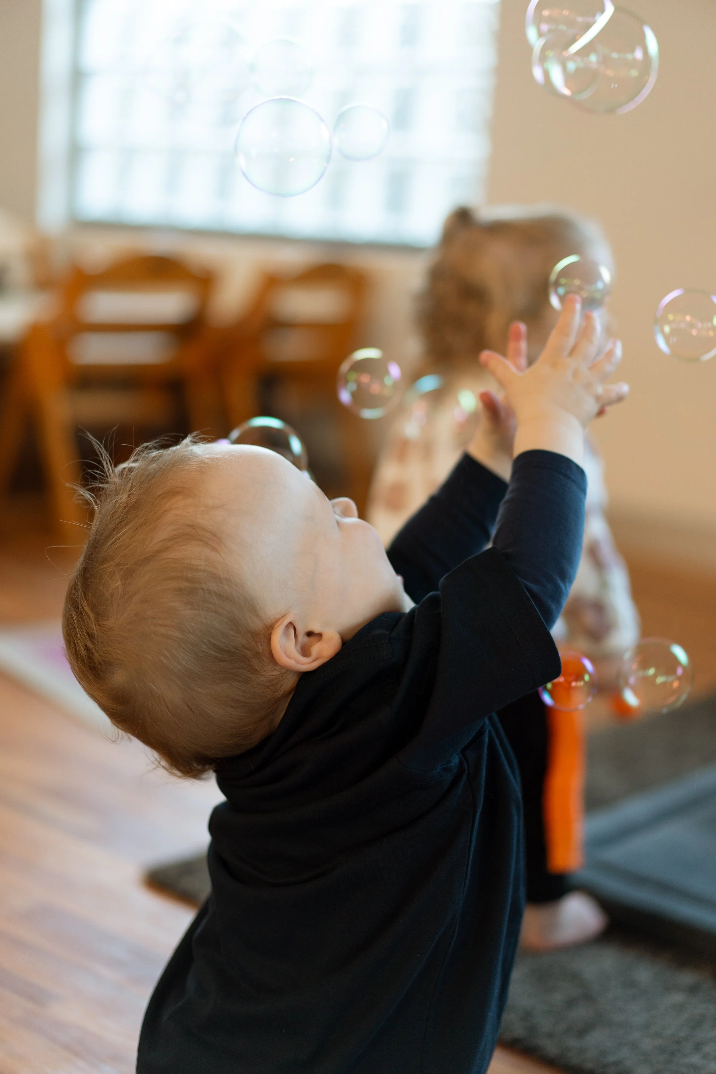 Child playing with floating soap bubbles in a room with wooden chairs and a window in the background.