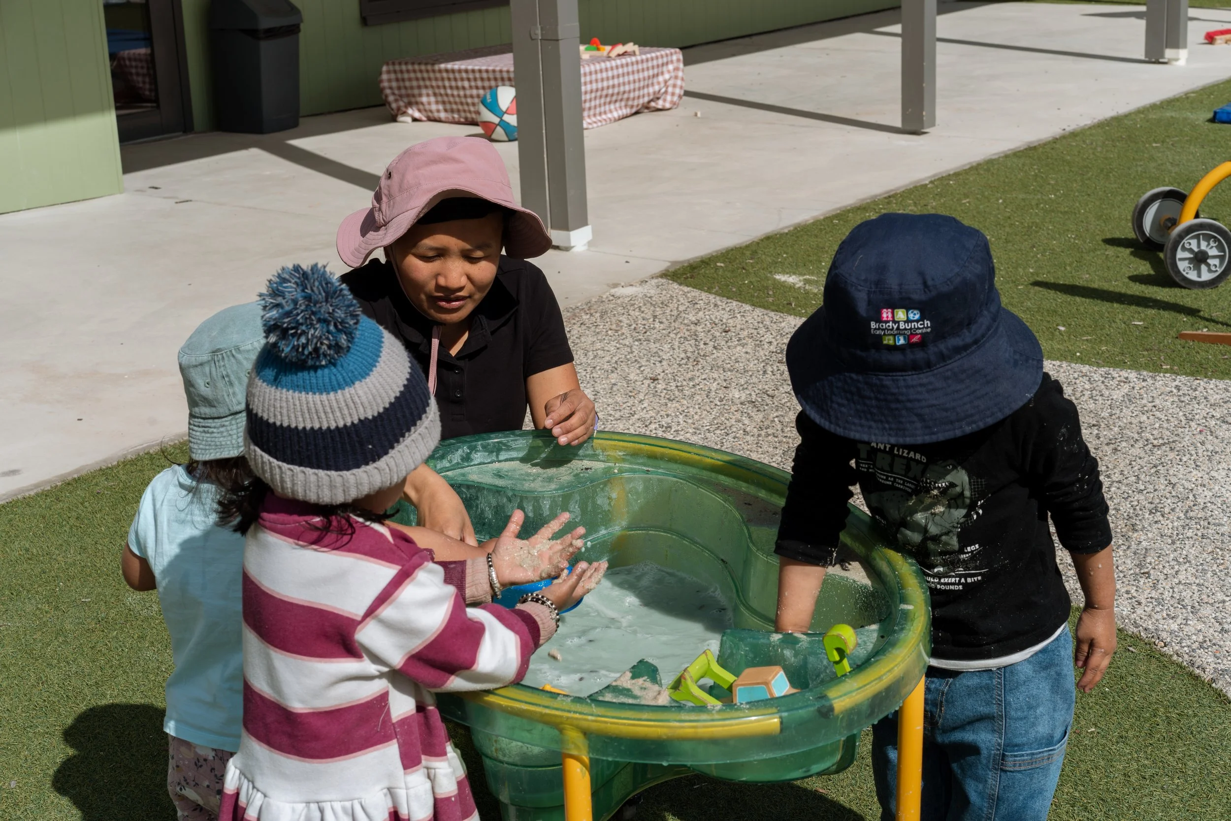 Children playing with water in a small pool outdoors, with an adult supervising. The setting appears to be a recreational area or playground near a building.