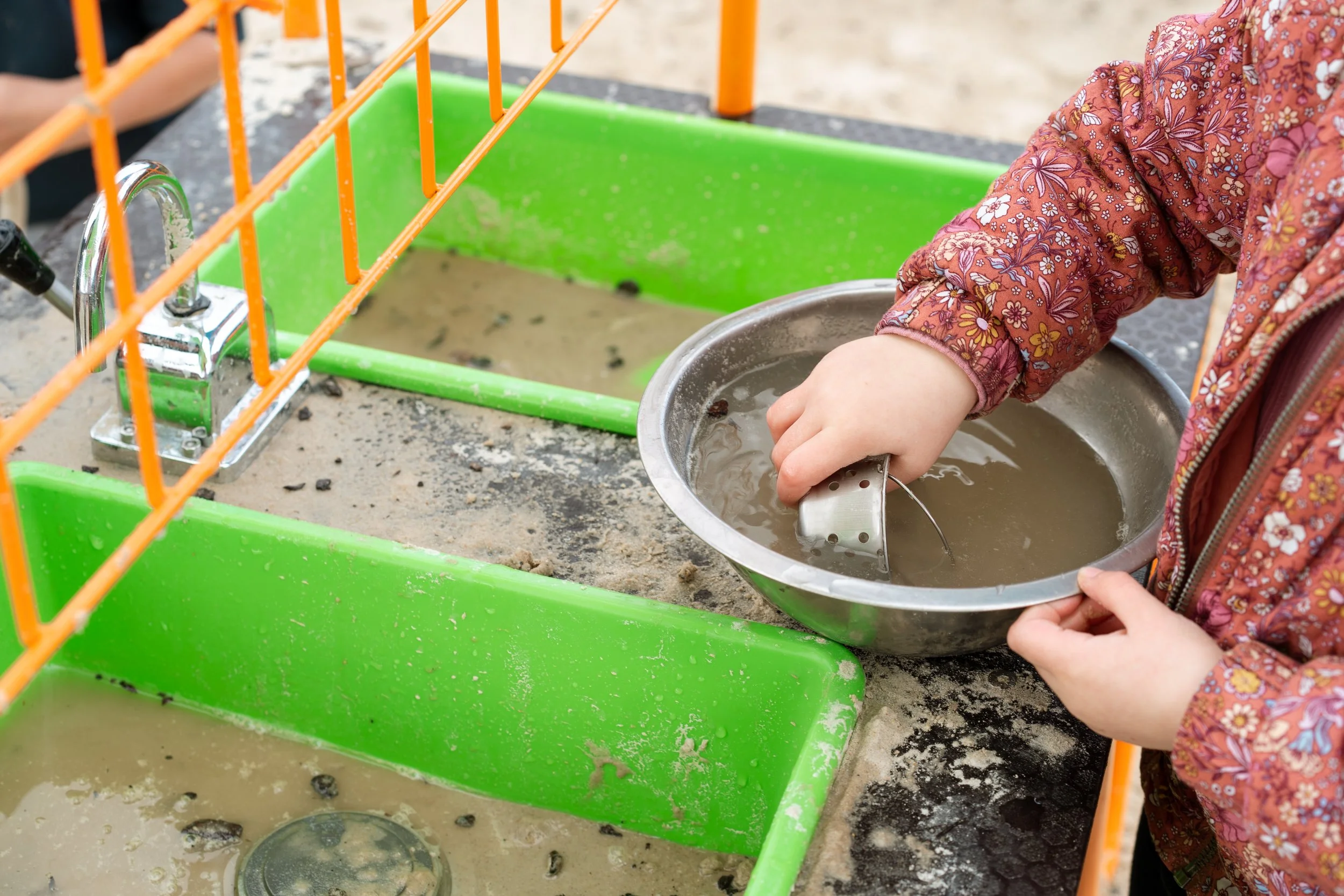 Child washing rocks in a metal bowl with muddy water, next to a green plastic sink filled with muddy water and small rocks.