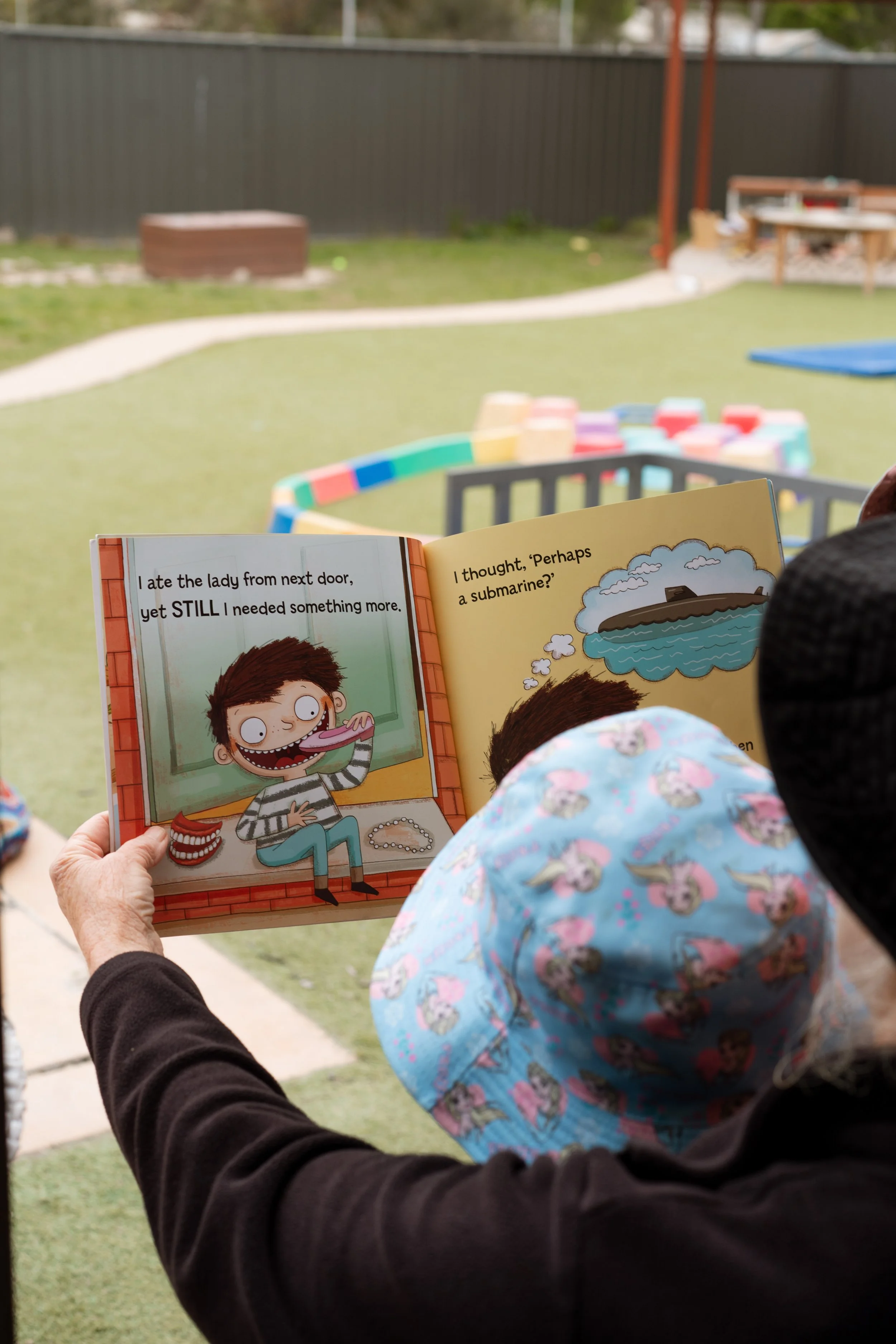 A person holding a children's book outdoors, with a child wearing a blue hat nearby. The scene includes a fenced yard with a sandbox and yard toys.