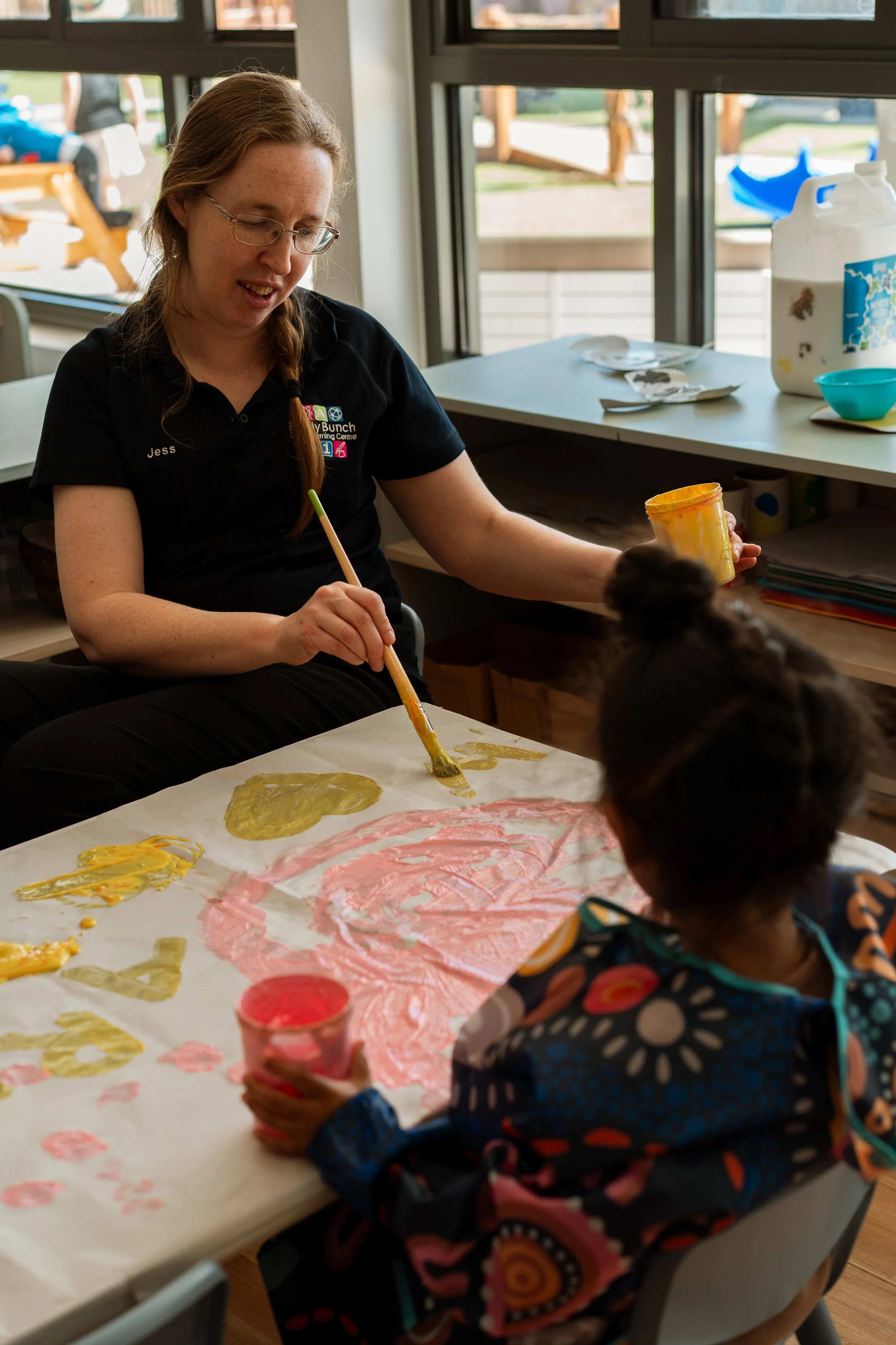 A woman with glasses and a ponytail painting on a large sheet of paper with children in a classroom or art room, with paint jars and art supplies on the table.