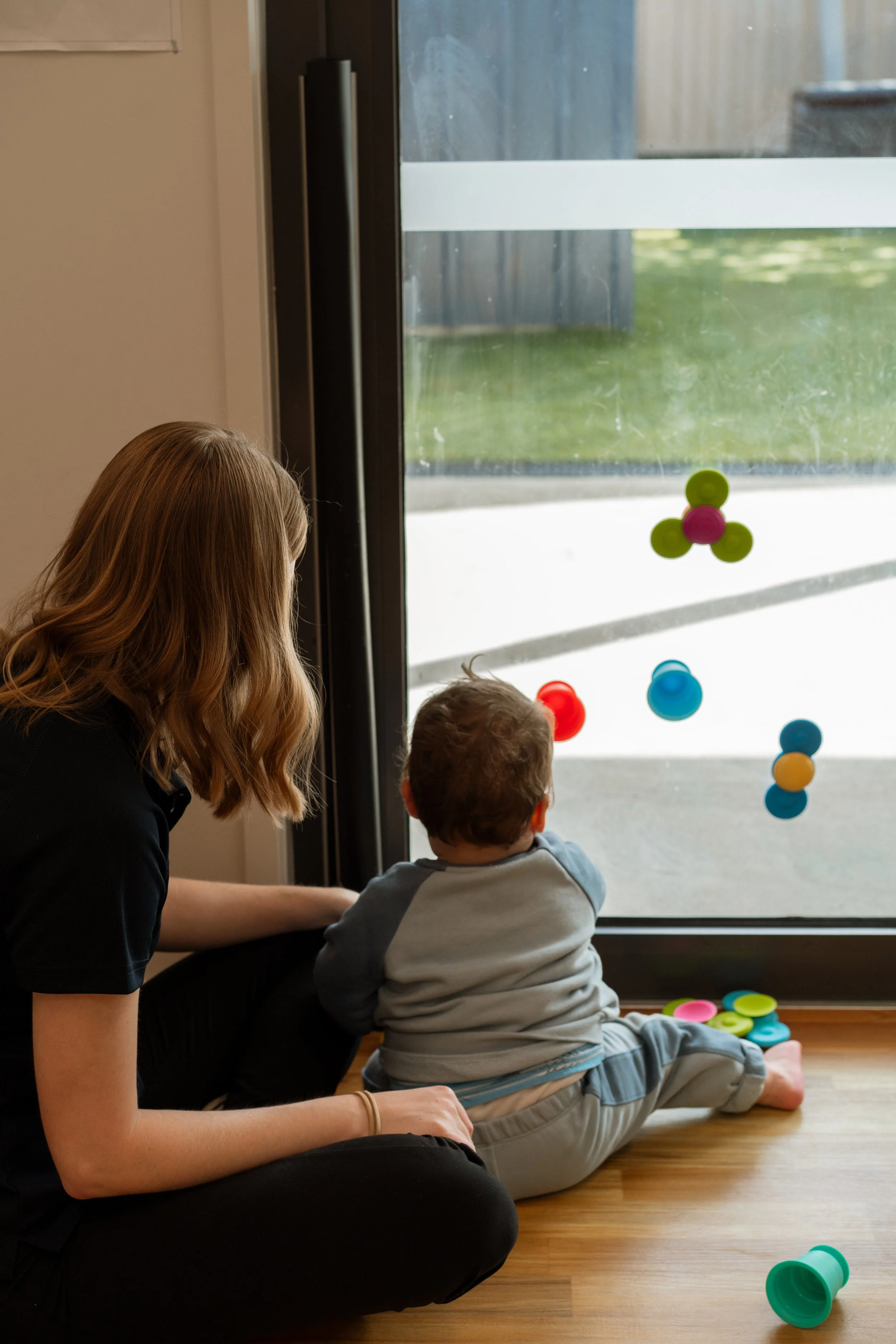 A woman and a young boy sitting on a wooden floor near a glass door, looking outside. The woman has long, wavy blonde hair, and the boy has short, dark hair. Colorful circular discs are scattered on the floor and attached to the glass door.