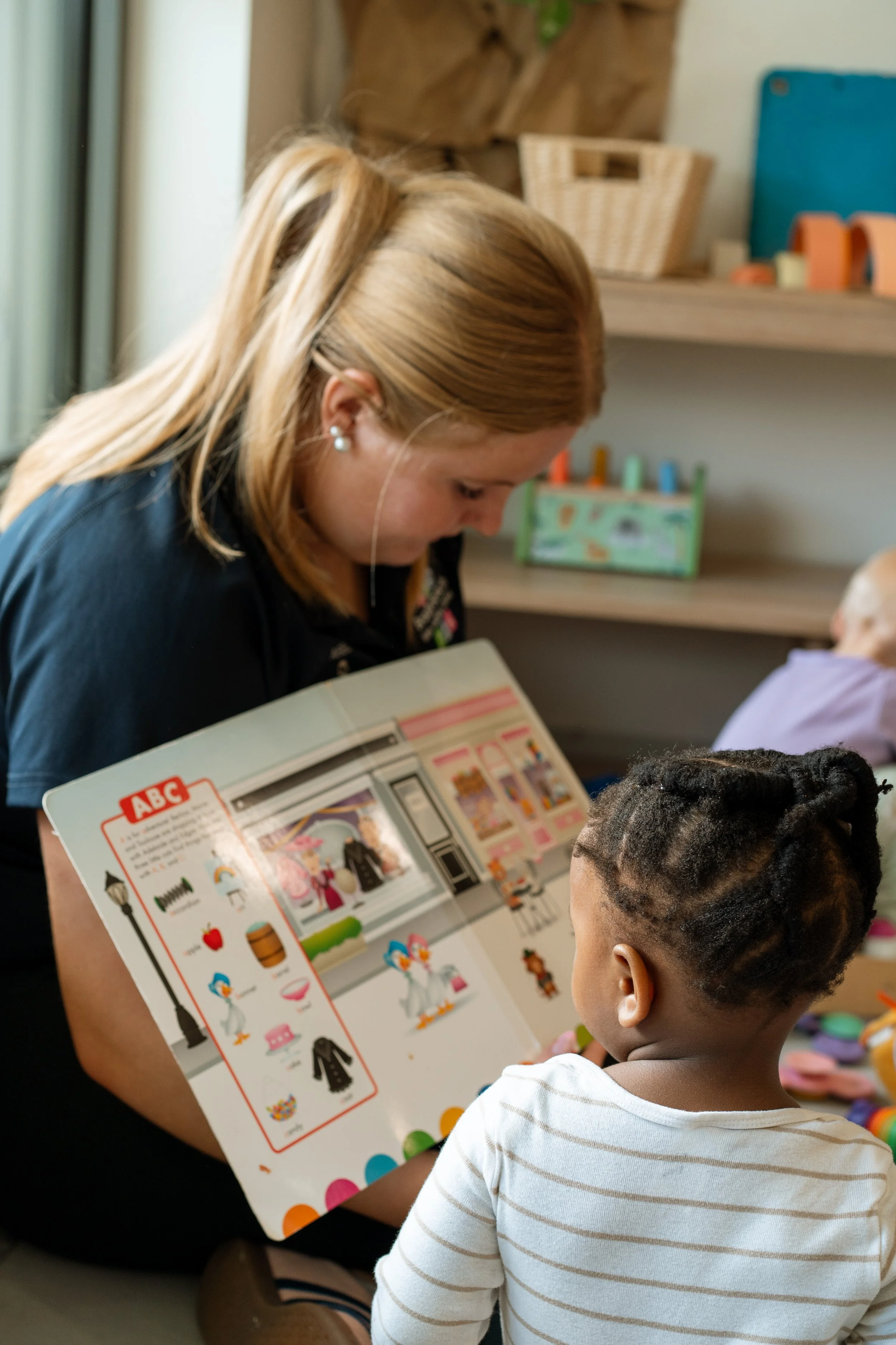A woman with long blonde hair and pearl earrings shows a colorful illustrated book to a young boy with braided hair and a striped white shirt in a cozy indoor setting.