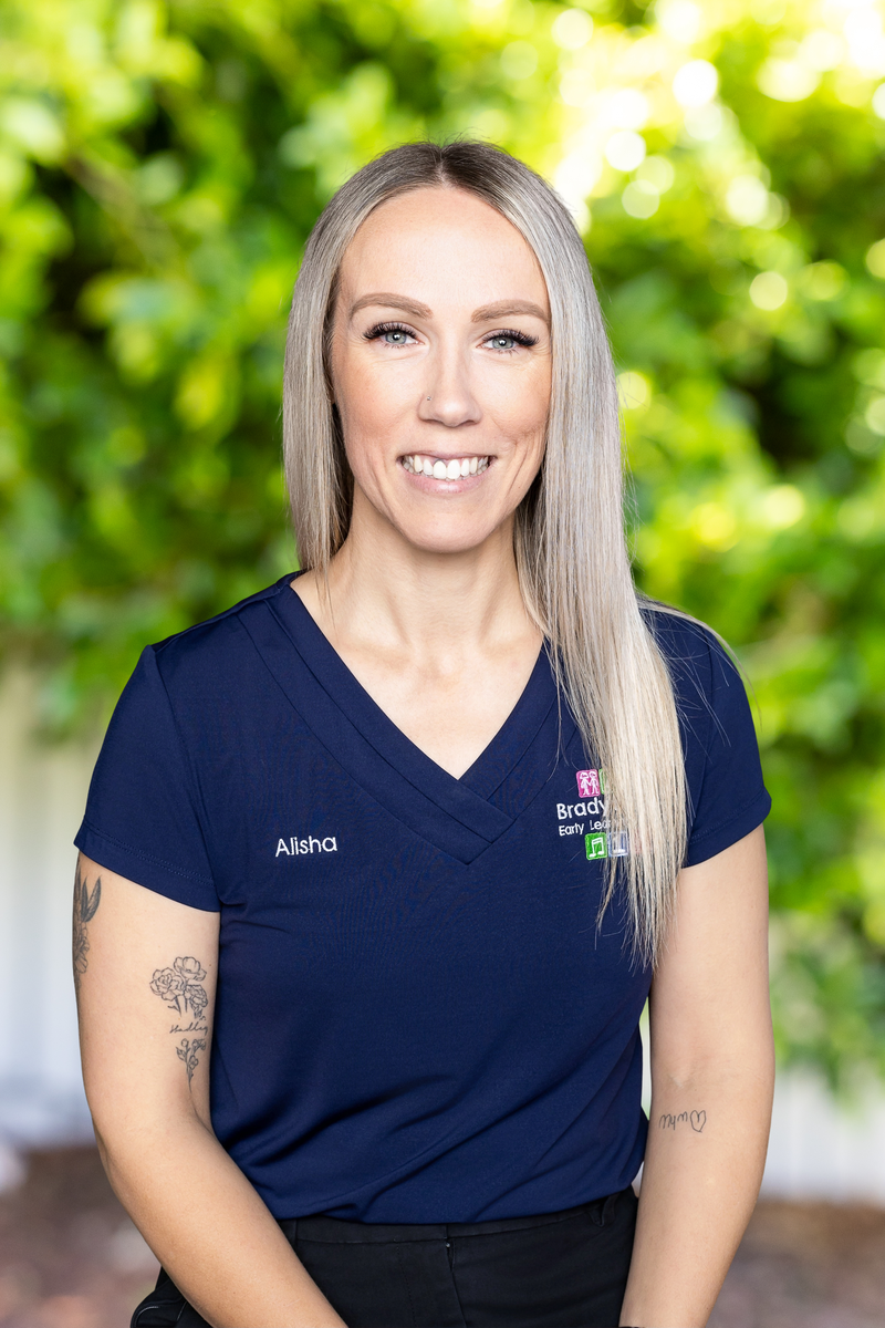 A woman with long, straight blonde hair and tattoos on her arms, wearing a navy blue scrub top with the name 'Alisha' embroidered on it, standing outdoors with a background of green trees and blurred sunlight.
