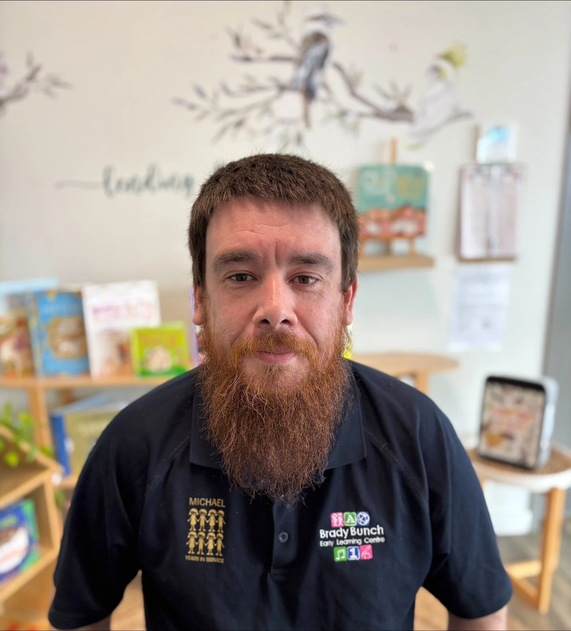 A man with short brown hair and a beard wearing a black shirt with embroidered text and various patches. The background shows a blurred classroom or childcare setting with books and educational materials on shelves.