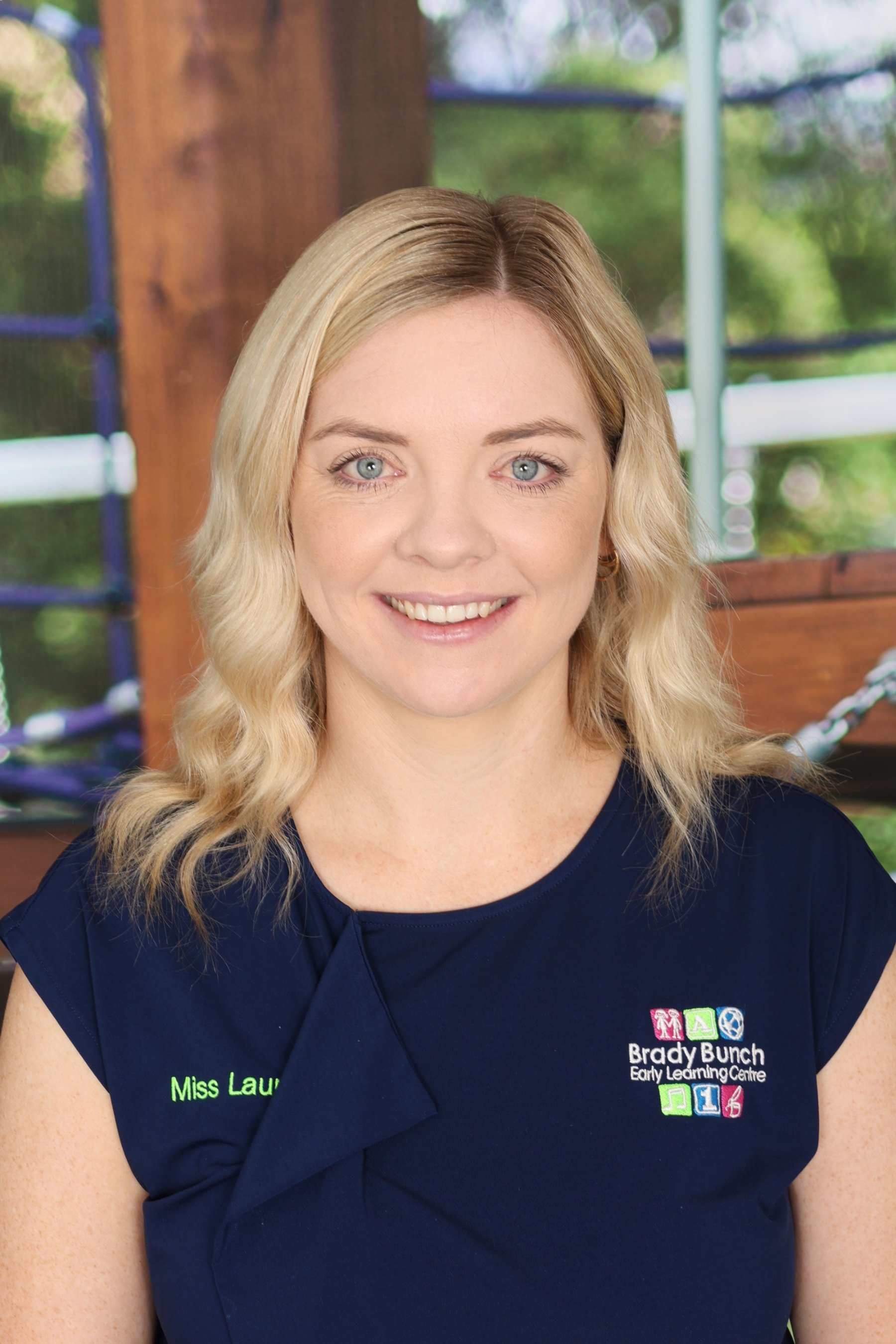 Close-up of a smiling woman with blonde hair in a navy uniform at Braydy Bunch Early Learning Centre with a colorful logo, standing outdoors with green trees and wooden and metal play structures in the background.