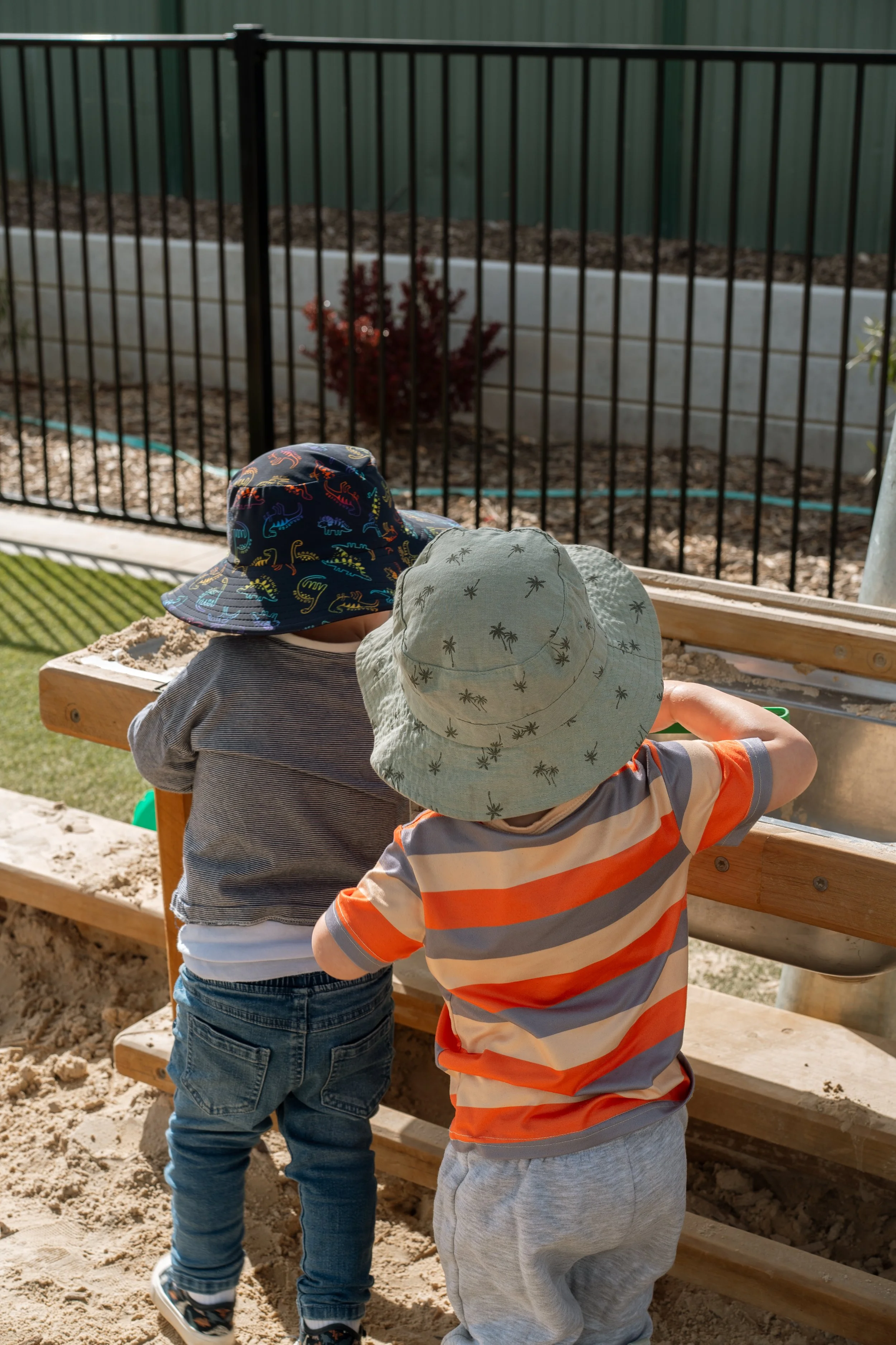 Two children wearing hats, standing by a sandbox at a playground, with a black fence and some plants in the background.