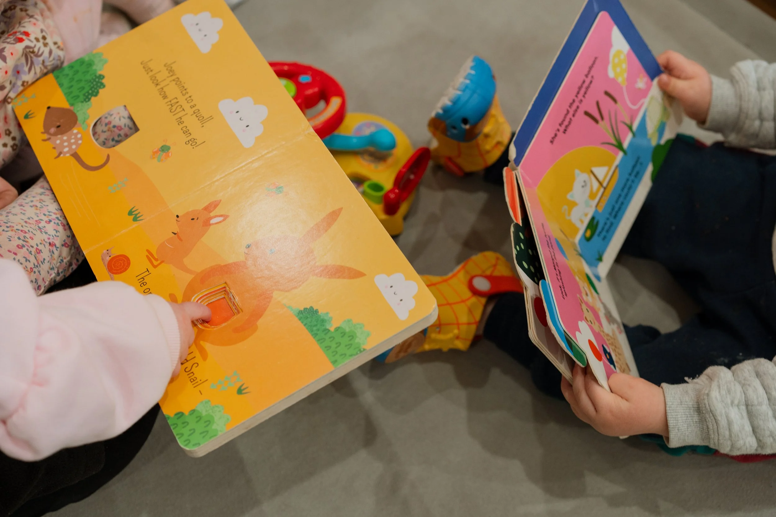 Six young children sitting on a rug in a classroom, smiling and looking at picture books. The background features wooden shelves, a decorative lattice with green leaves, and artwork, creating a cheerful learning environment.