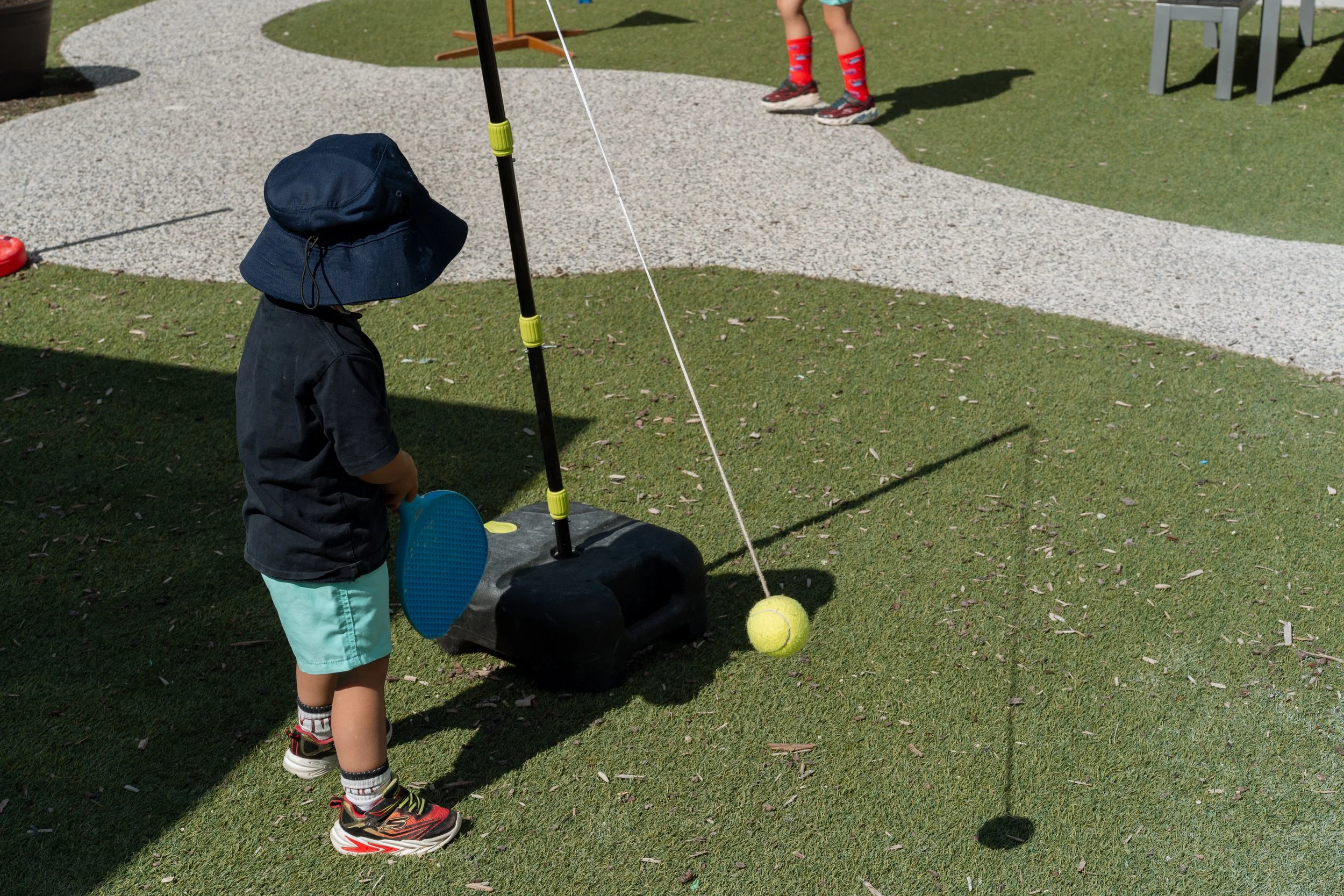 A young child wearing a white sun hat and a black T-shirt is sitting at the top of a green slide on a playground, smiling. The playground is outdoors with green grass, trees, and a blue canopy overhead.