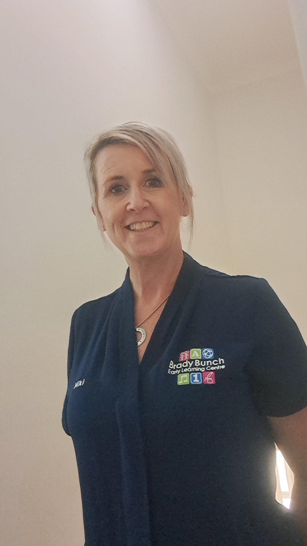 A smiling woman with short blonde hair wearing a navy blue shirt with colorful logo embroidery representing Brady Bunch Early Learning Center, and a necklace, standing against a plain off-white wall.
