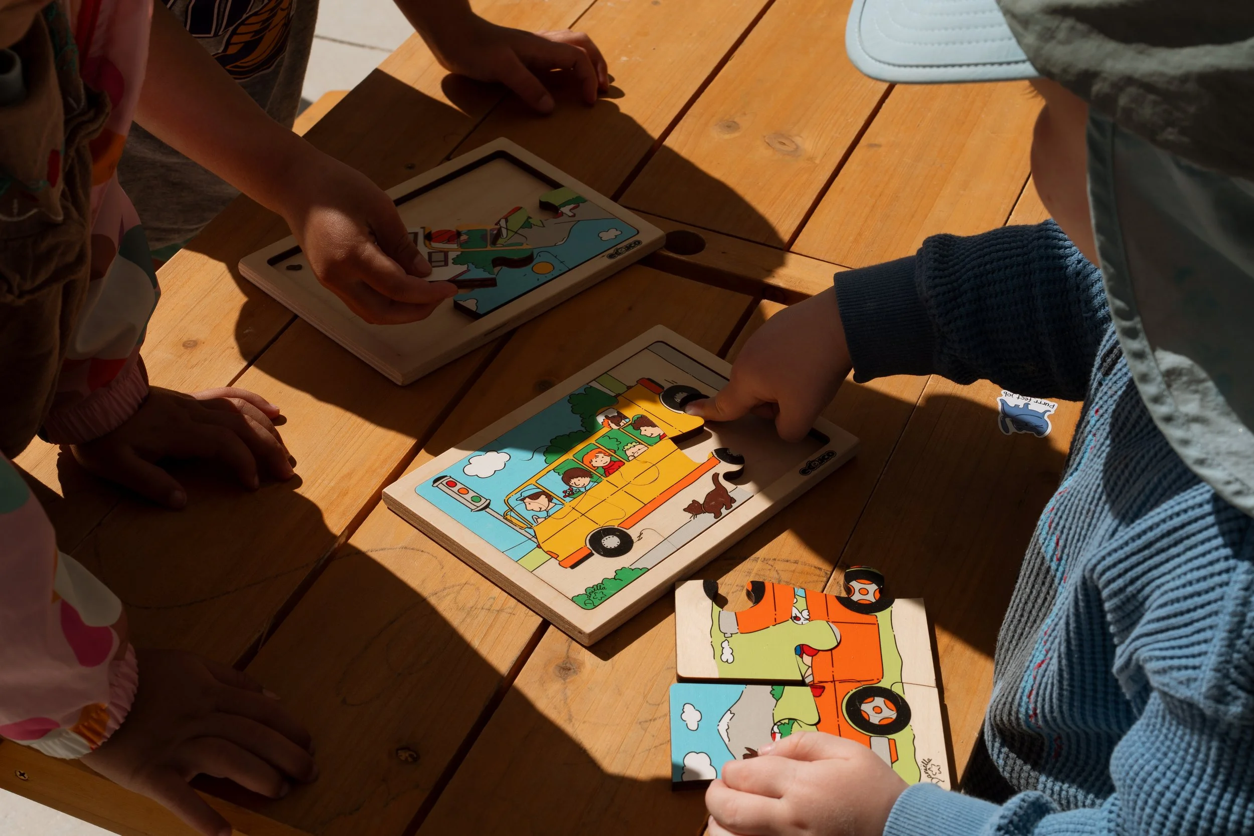 A group of young children sitting around a table playing with toy construction vehicles in a well-lit classroom. The room has white walls, wooden furniture, and various educational displays.