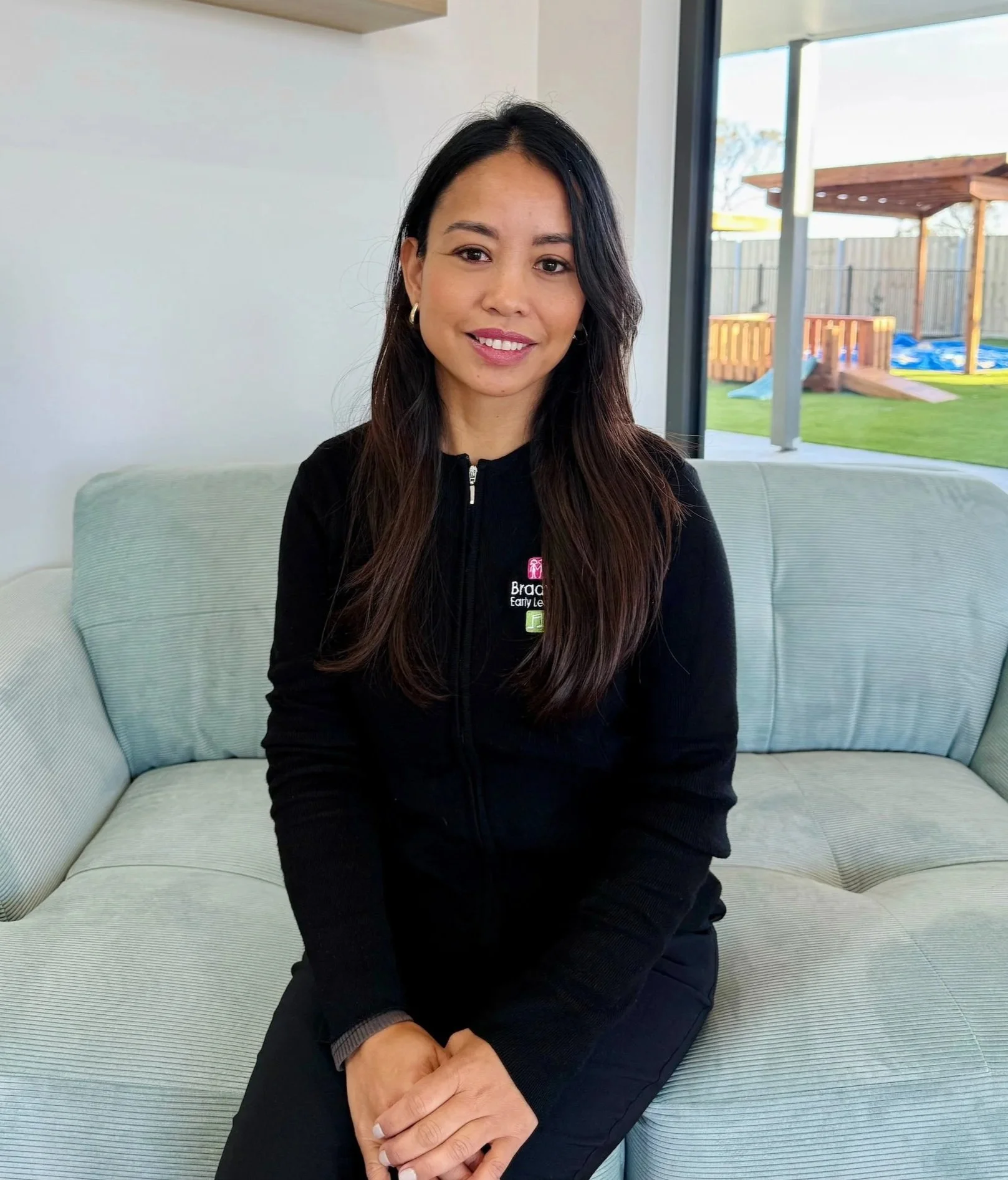 A woman sitting on a light-colored sofa in a bright room, smiling at the camera, with a large window revealing a backyard with a wooden play structure and a blue pool.